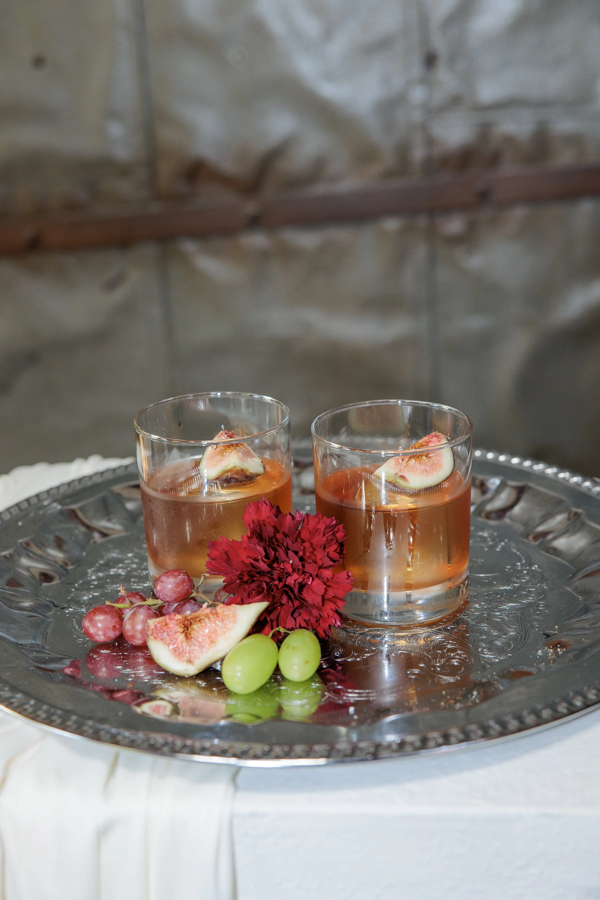 Two glasses of rose-colored drink with fig slices inside, on a silver tray with grapes and a red flower, against a textured gray wall background.