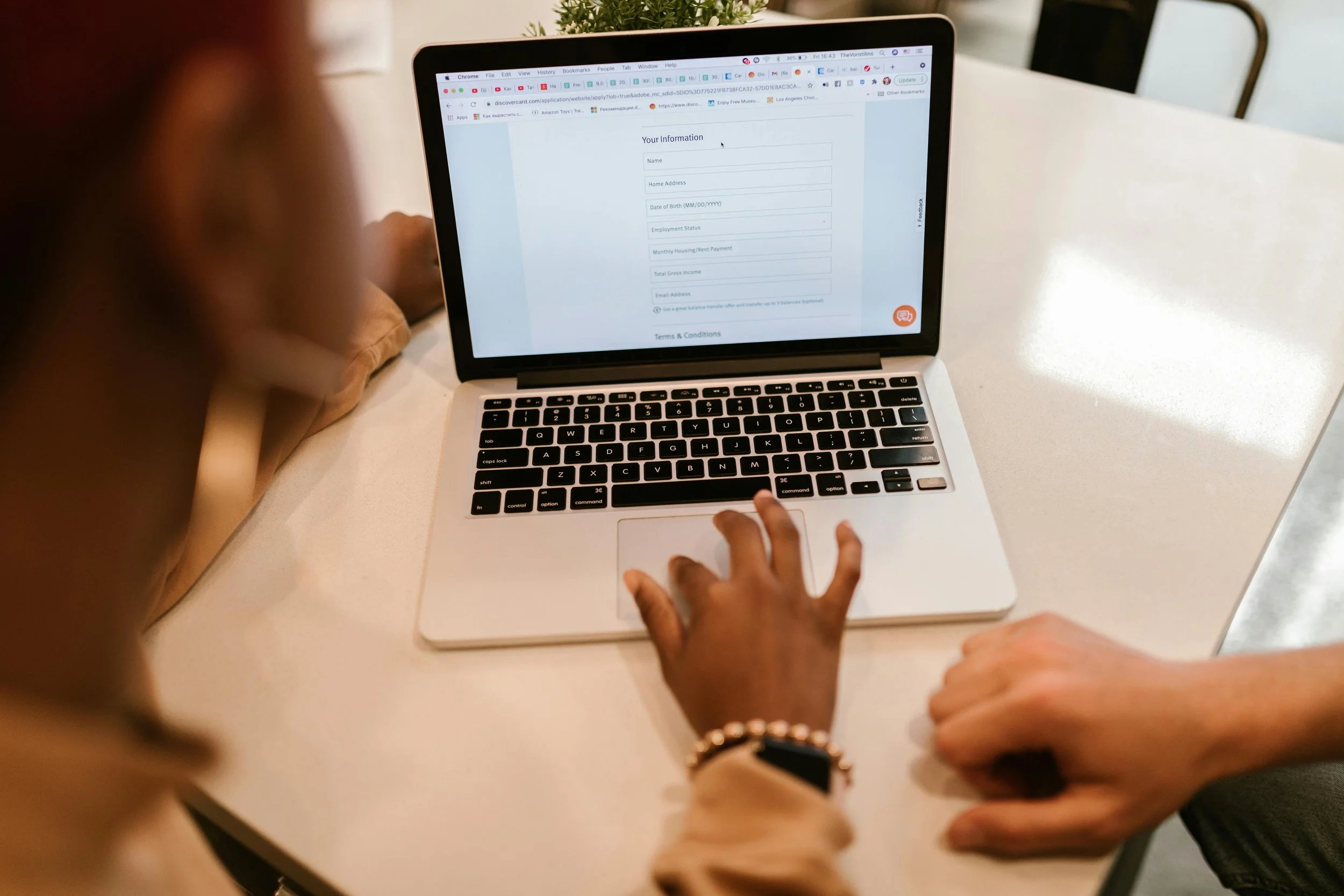 Person using a laptop at a white table, filling out an online form on the screen, with another person's arm resting on the table.