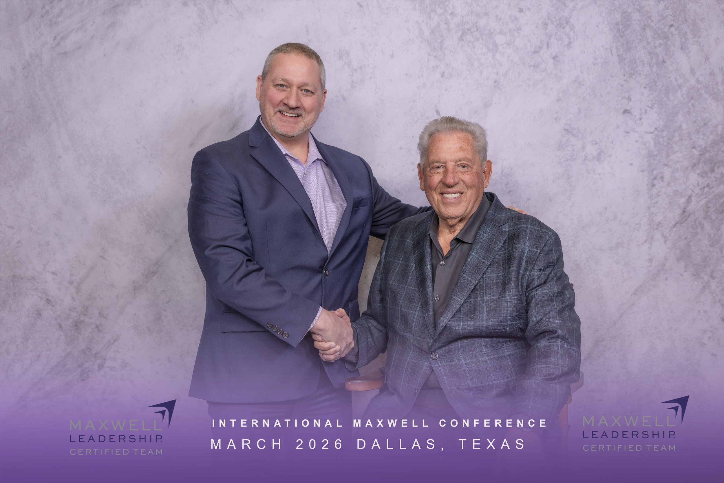 Two men shaking hands, one standing and one sitting, at a conference in Dallas, Texas, in March 2026. They are dressed in business suits, smiling, with a gray textured background. There is text on the image that reads "International Maxwell Conference, March 2026 Dallas Texas" and inclusive of Maxwell Leadership Certified Team logos.
