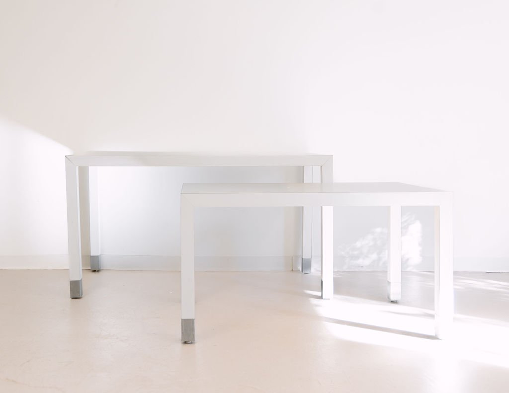 Two minimalist white tables against a white wall in a bright room.