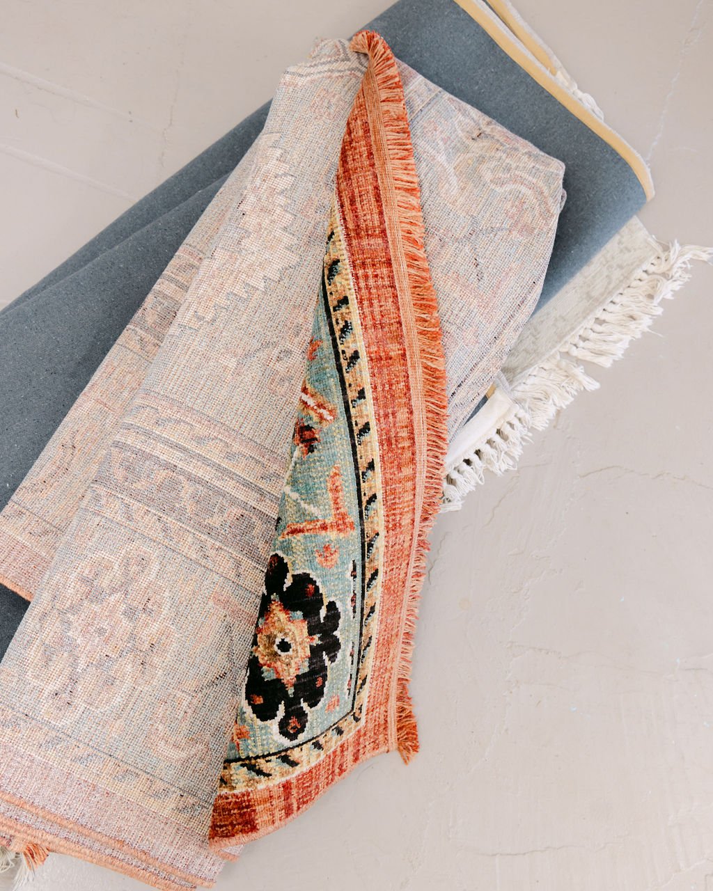 A stack of rolled area rugs with various intricate patterns and colors, including a visible section with a floral motif and fringed edges.