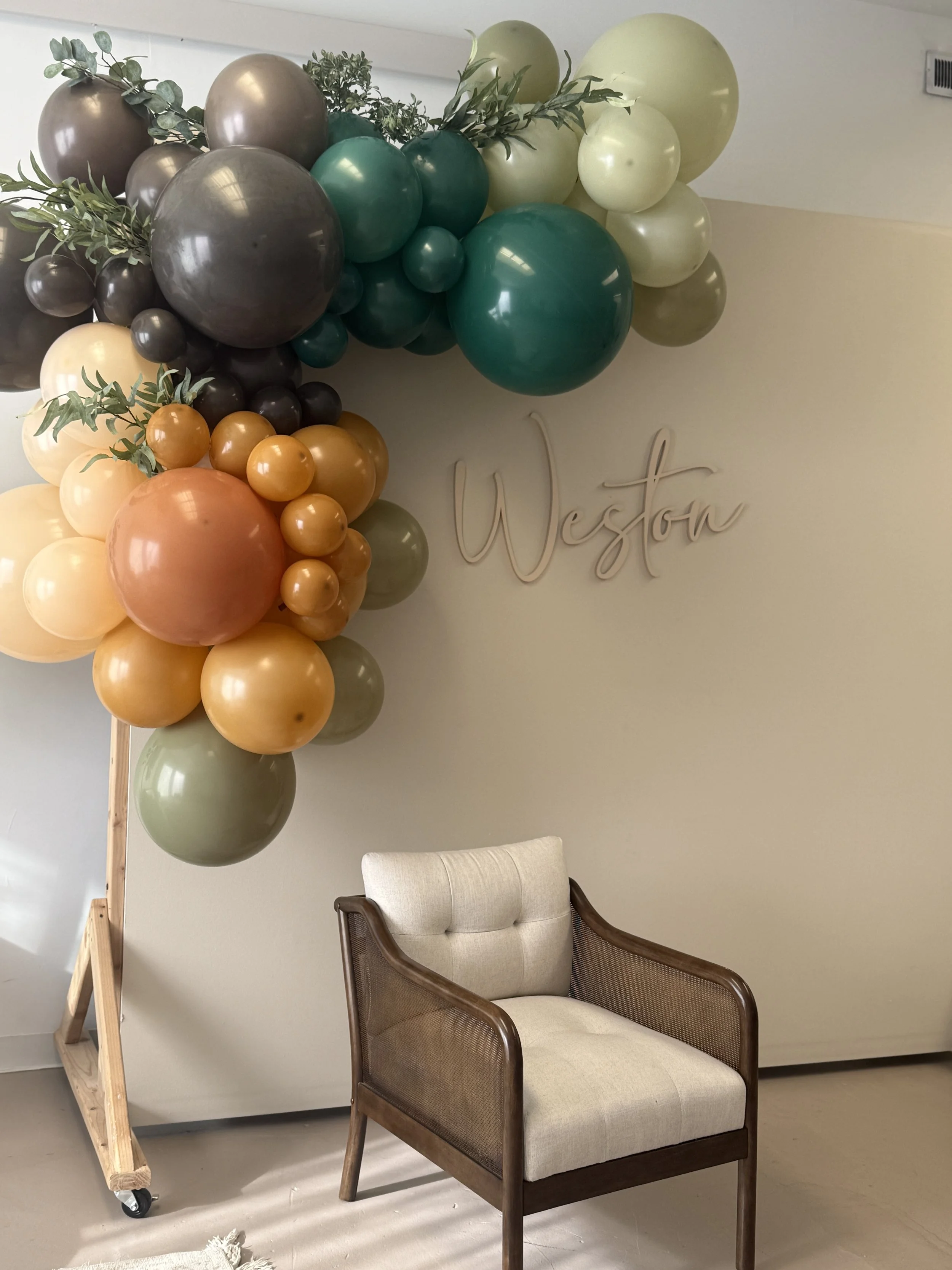 Woman sitting on a beige couch, holding a drink, next to a decorative balloon display with white and beige balloons. Wooden table with a bottle in an ice bucket and a plate of cookies. Clear chair in the foreground.