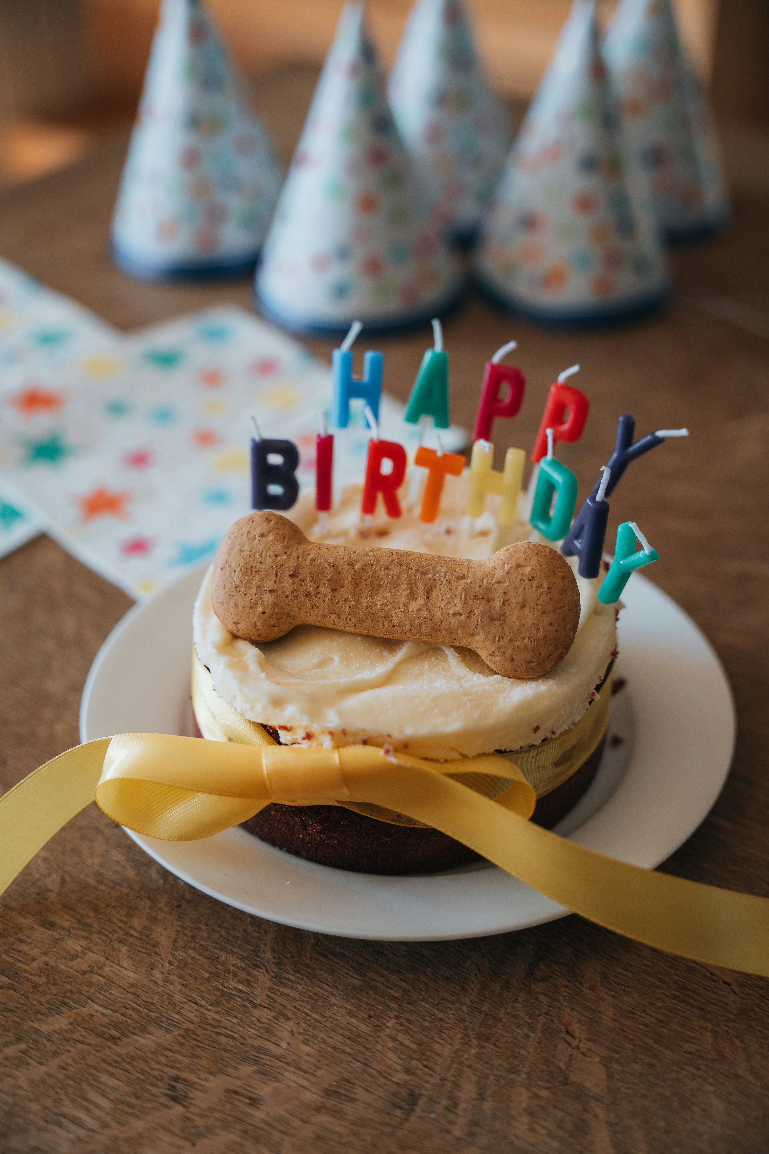 Close-up of a birthday cake with colorful candles spelling "HAPPY BIRTHDAY" and a dog-shaped cookie on top, surrounded by party hats and festive decorations.