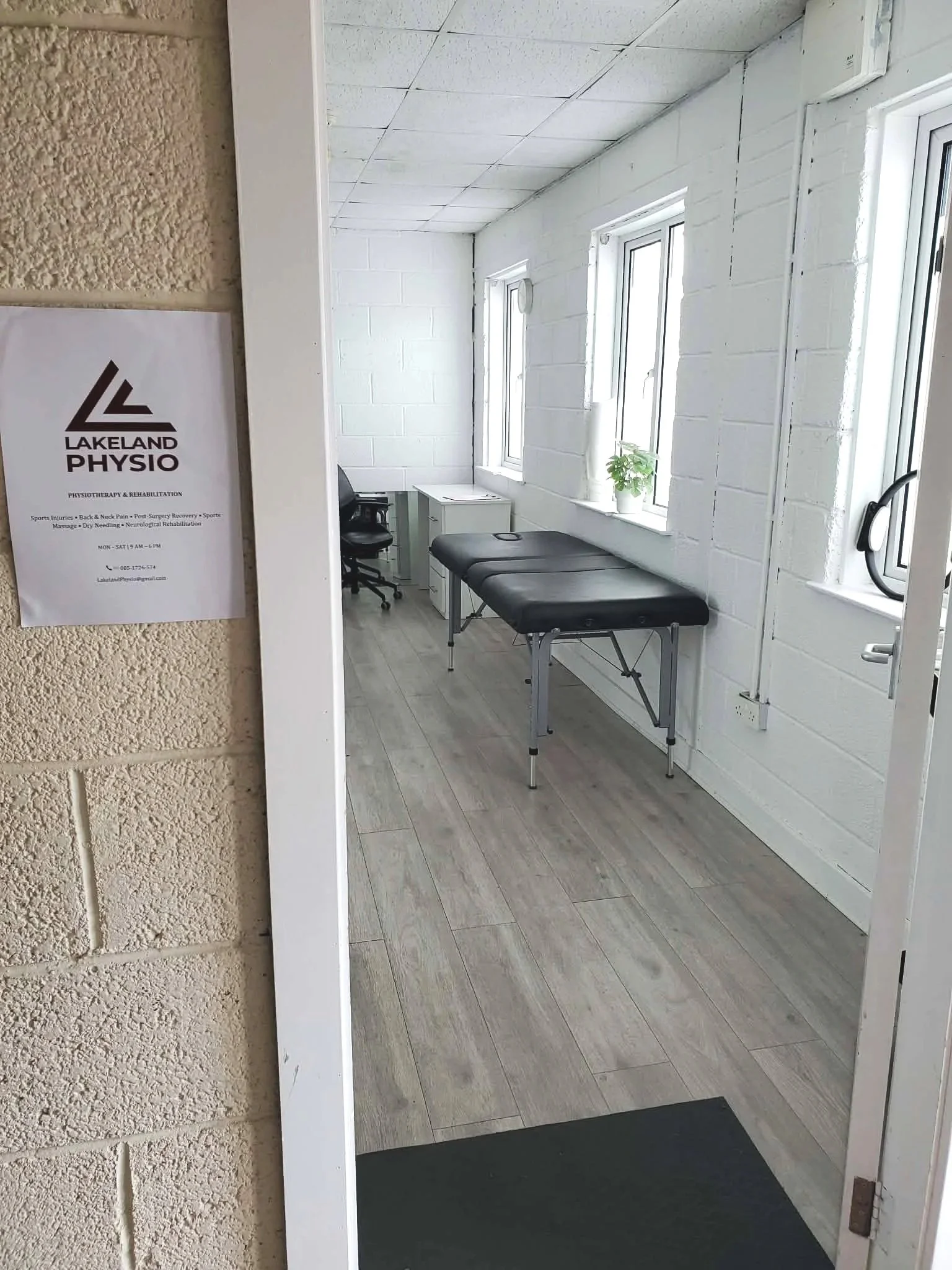 View into physical therapy clinic room with a black examination table, white cabinet, two windows with white frames, a potted plant, and an office chair.