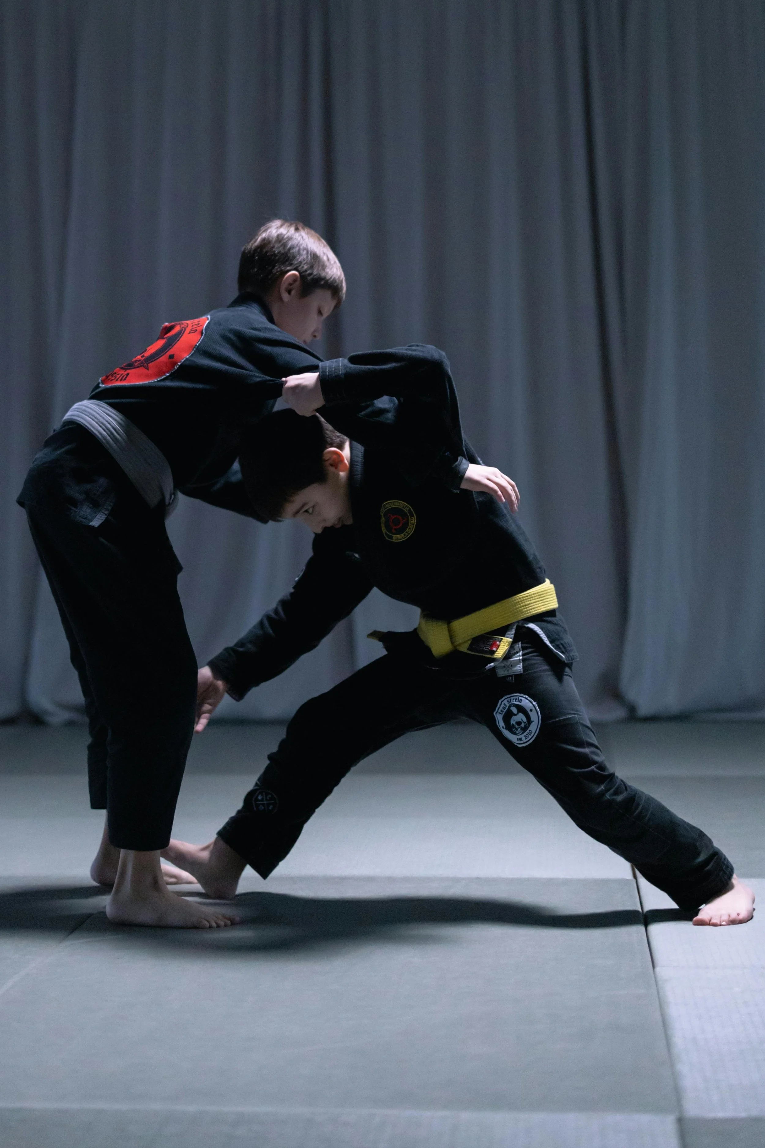 Two people practicing Brazilian Jiu-Jitsu in a martial arts gym. One person is standing and gripping the other, who is crouched with a yellow belt, on a grey mat with a curtain in the background.