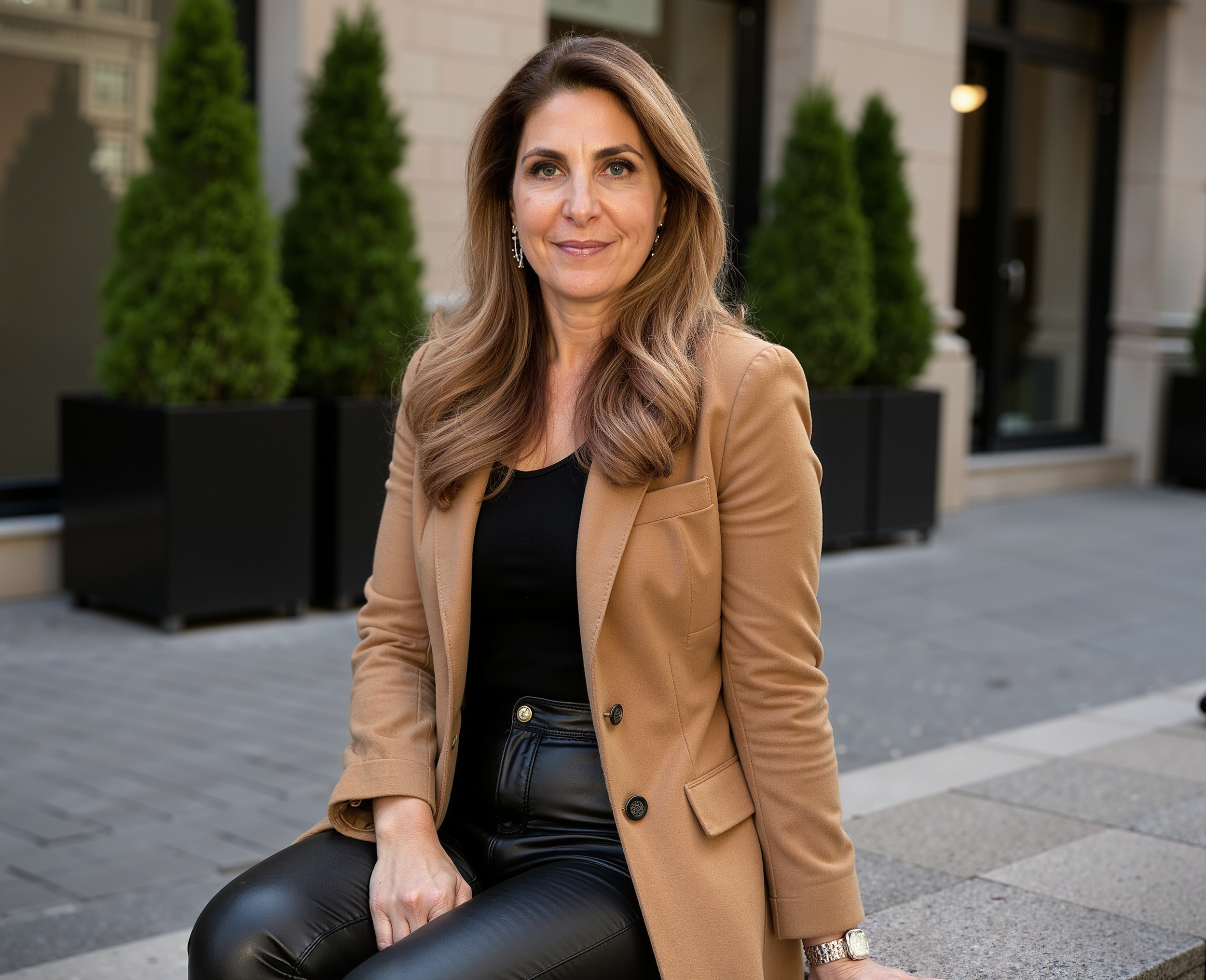 A woman sitting outdoors on a city sidewalk, wearing a tan blazer, black shirt, and black leather pants, with potted evergreen trees and a building behind her.
