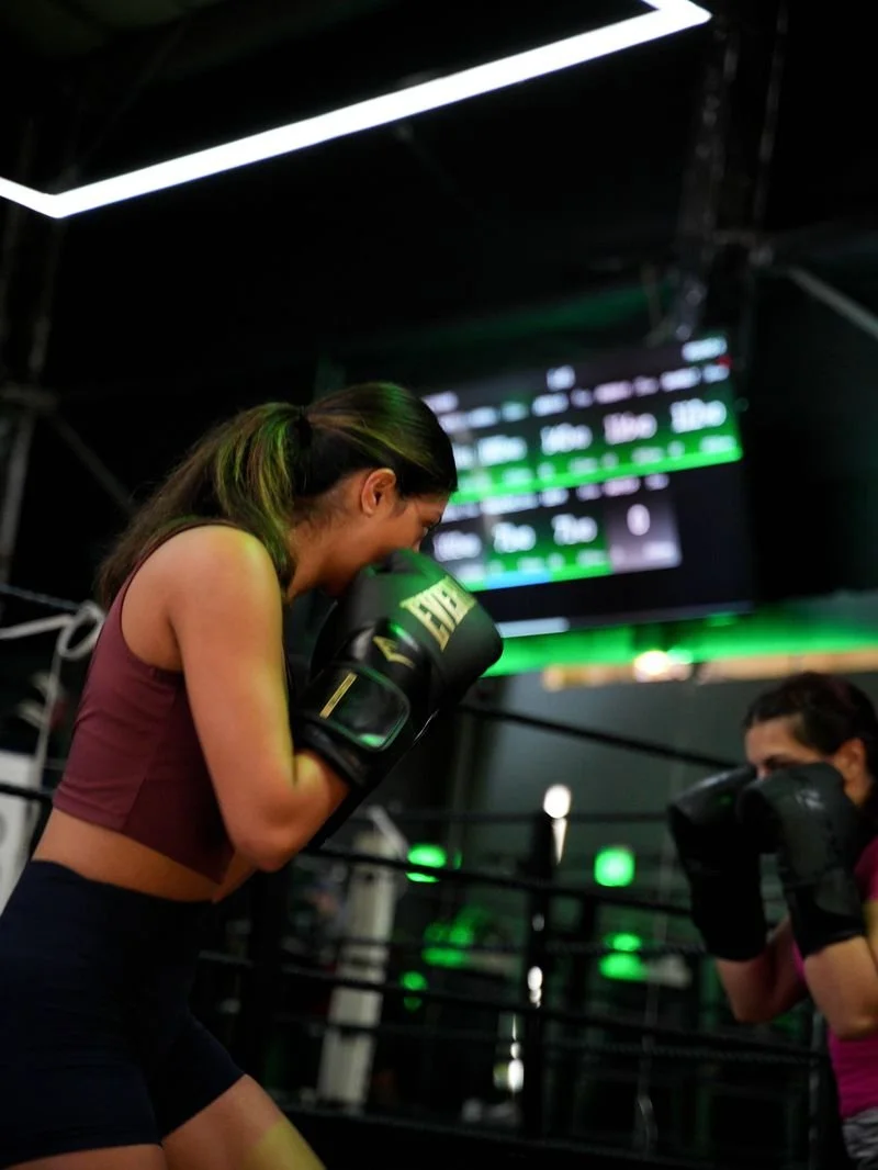 Two women boxing in a gym, one woman is wearing black gloves and a purple sports bra, and the other is in black gloves and a pink top. The woman in the foreground appears to be smiling or laughing.