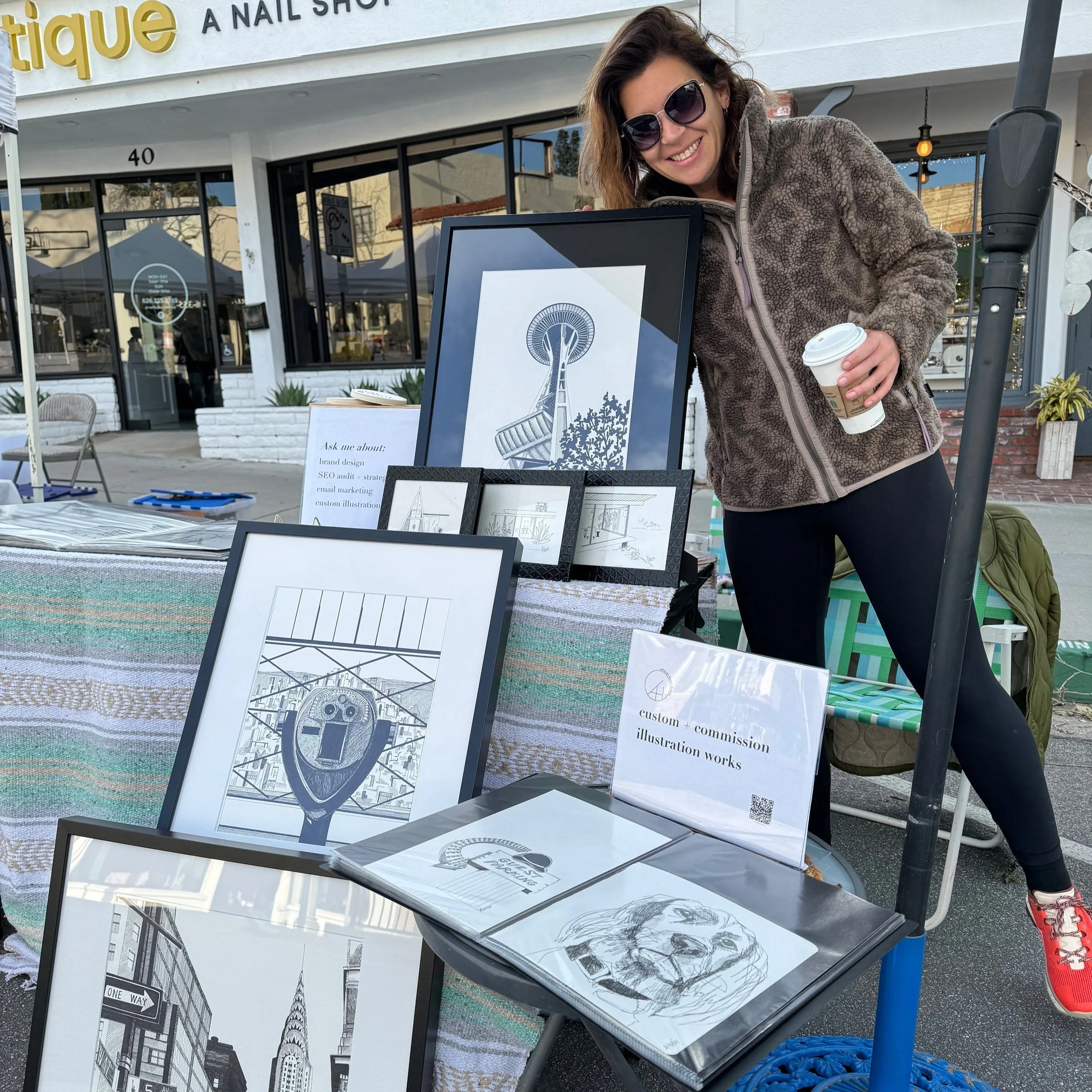 A smiling woman with sunglasses holding a coffee cup, standing next to an outdoor art booth showcasing framed illustrations and prints, with a building and store sign in the background.