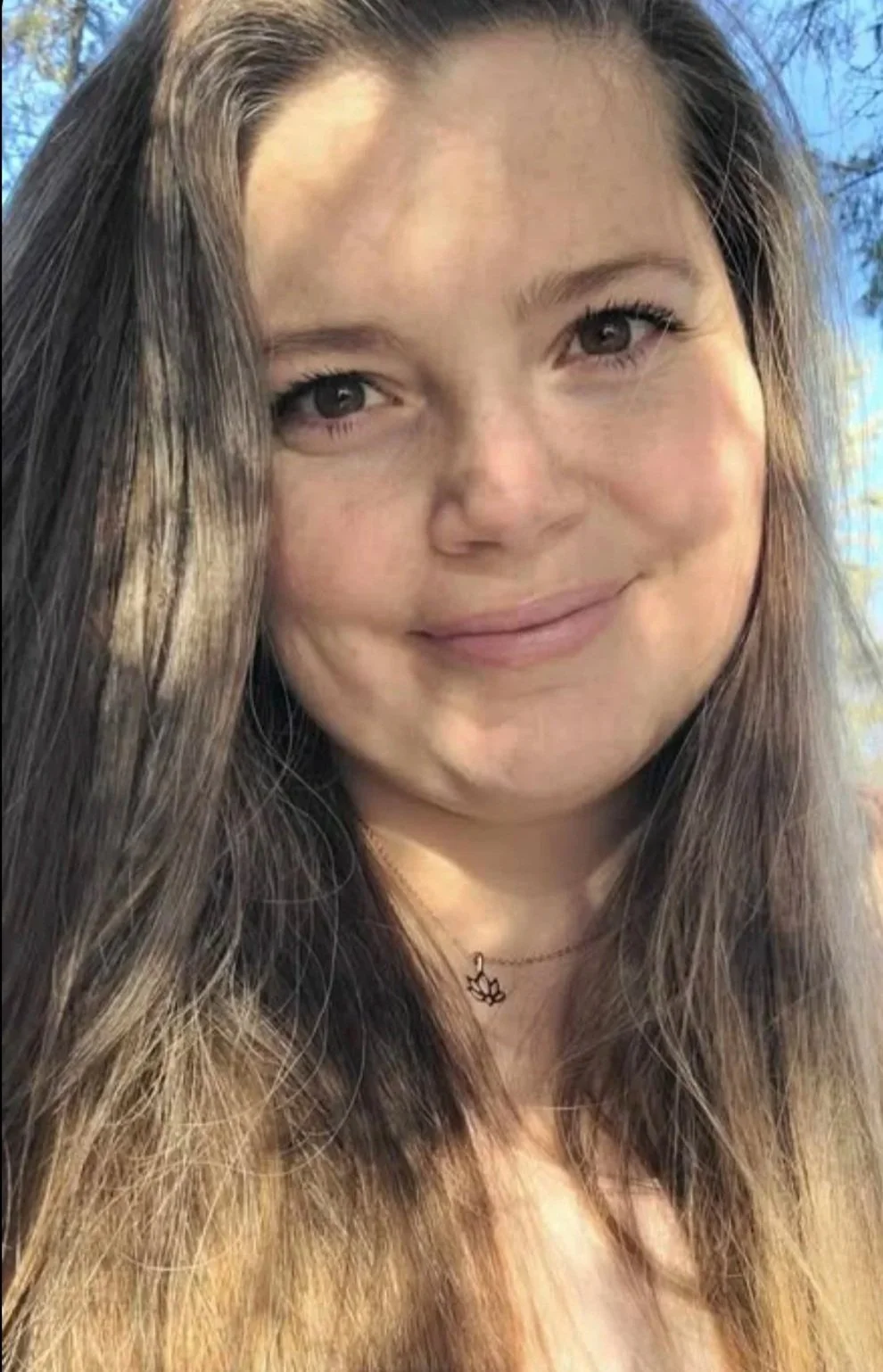 Close-up of a smiling woman with long brown hair, wearing a small necklace, outdoors with trees and blue sky in the background.