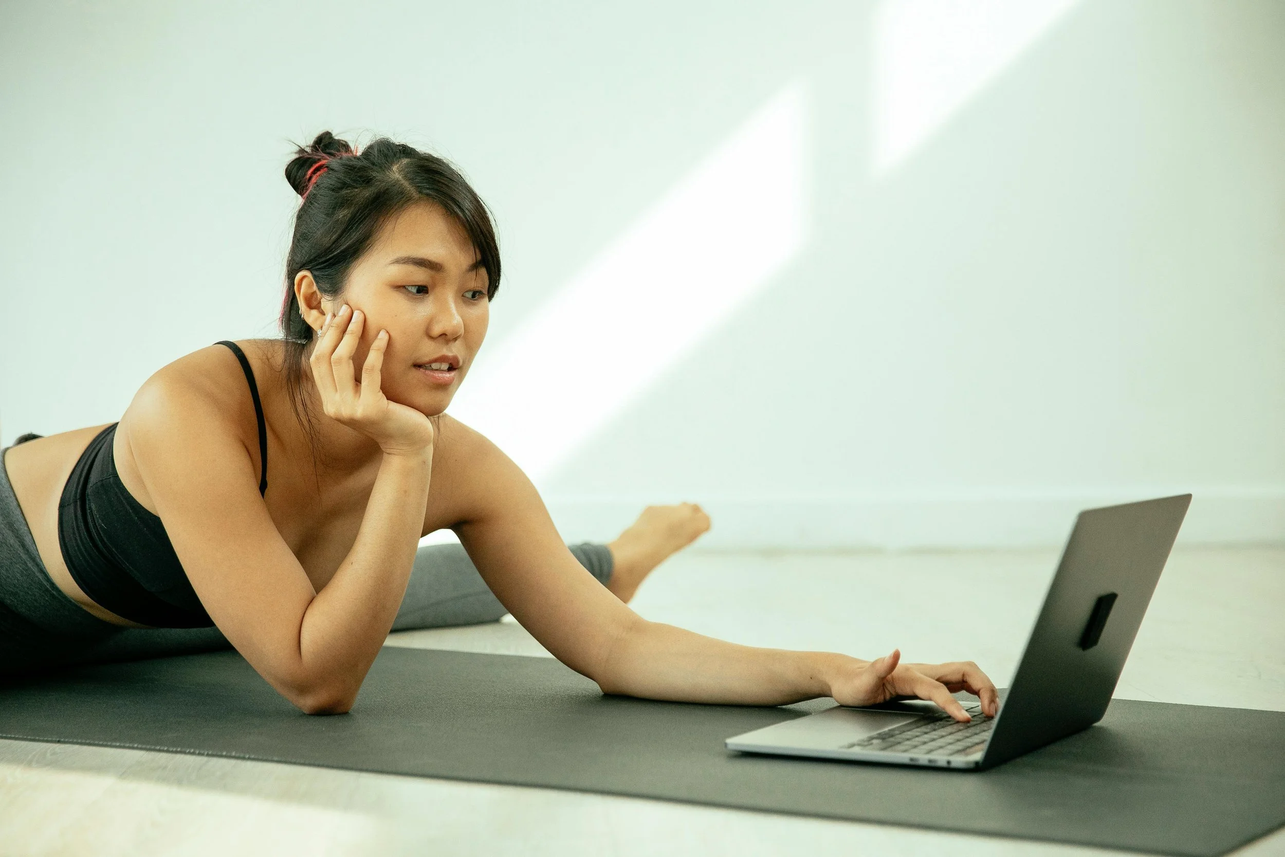 A woman lying on her stomach on a yoga mat using a laptop, with her chin resting on her hand, in a bright room.