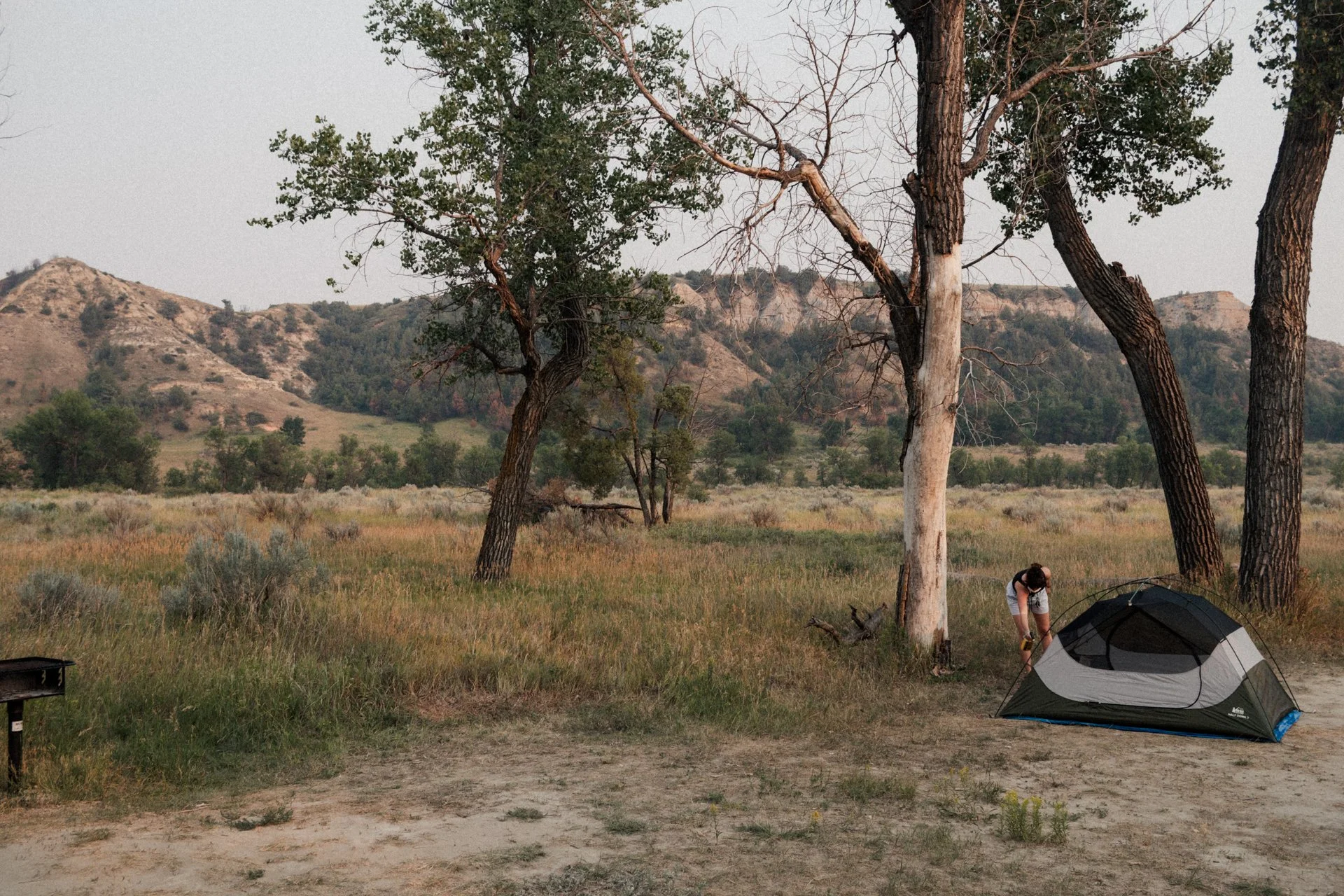 Person setting up a tent in a grassy field with trees and hills in the background.