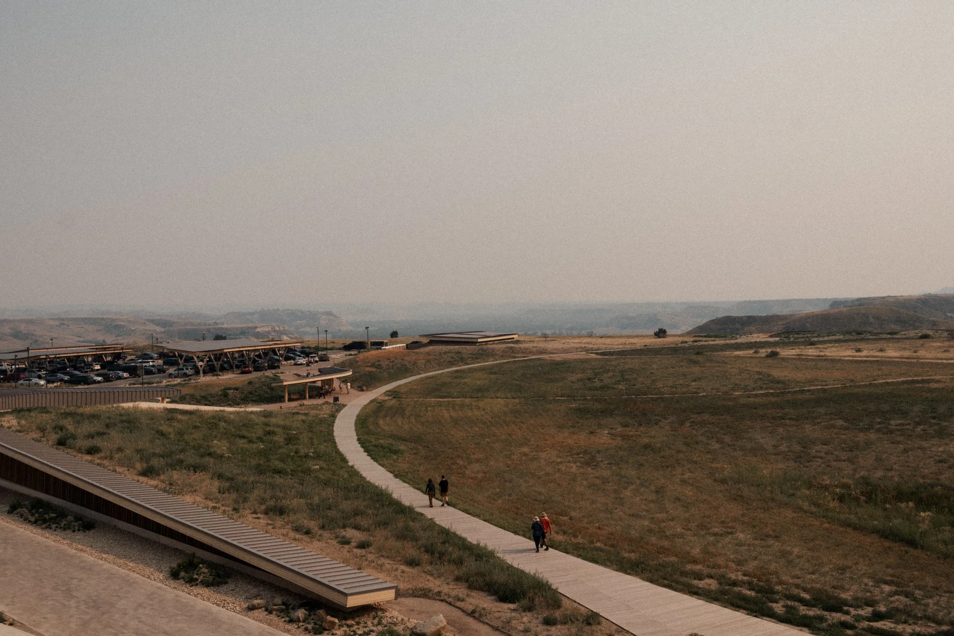 A scenic view of a park with a winding pathway, grassy fields, and a parking lot with cars. There are a few people walking along the path and a parking lot structure in the distance under a hazy sky.