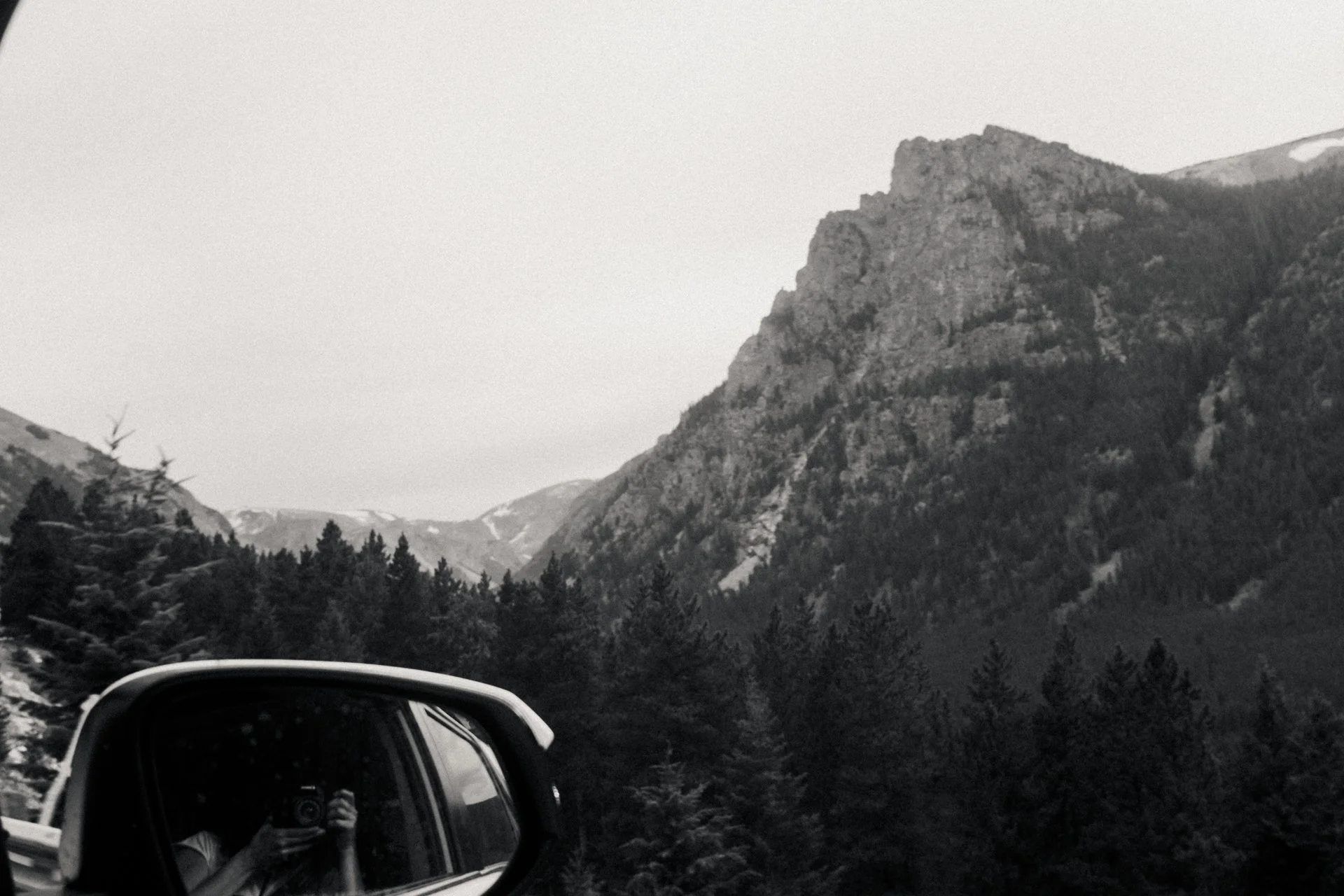 Black and white photo of tall mountain with rocky slopes, evergreen trees in foreground, and snow patches at the top, seen through the side mirror of a car.