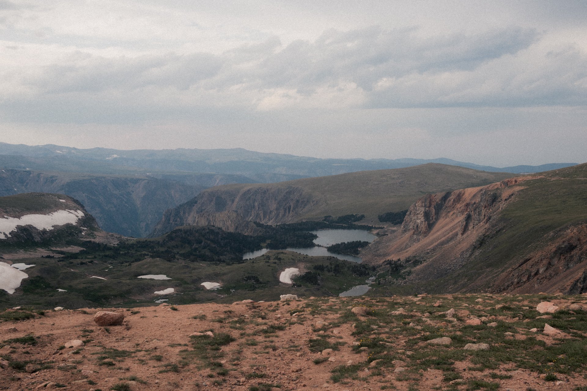 A rugged mountain landscape with cliffs, snow patches, a small lake, and rolling hills under a cloudy sky.
