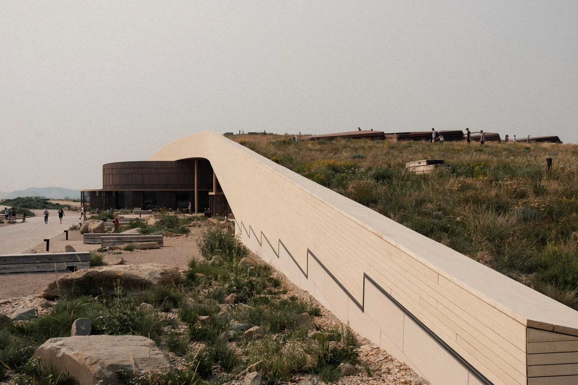 Contemporary building with curved roof and walkway, with people walking on grassy hillside and outdoor plaza area, under overcast sky.