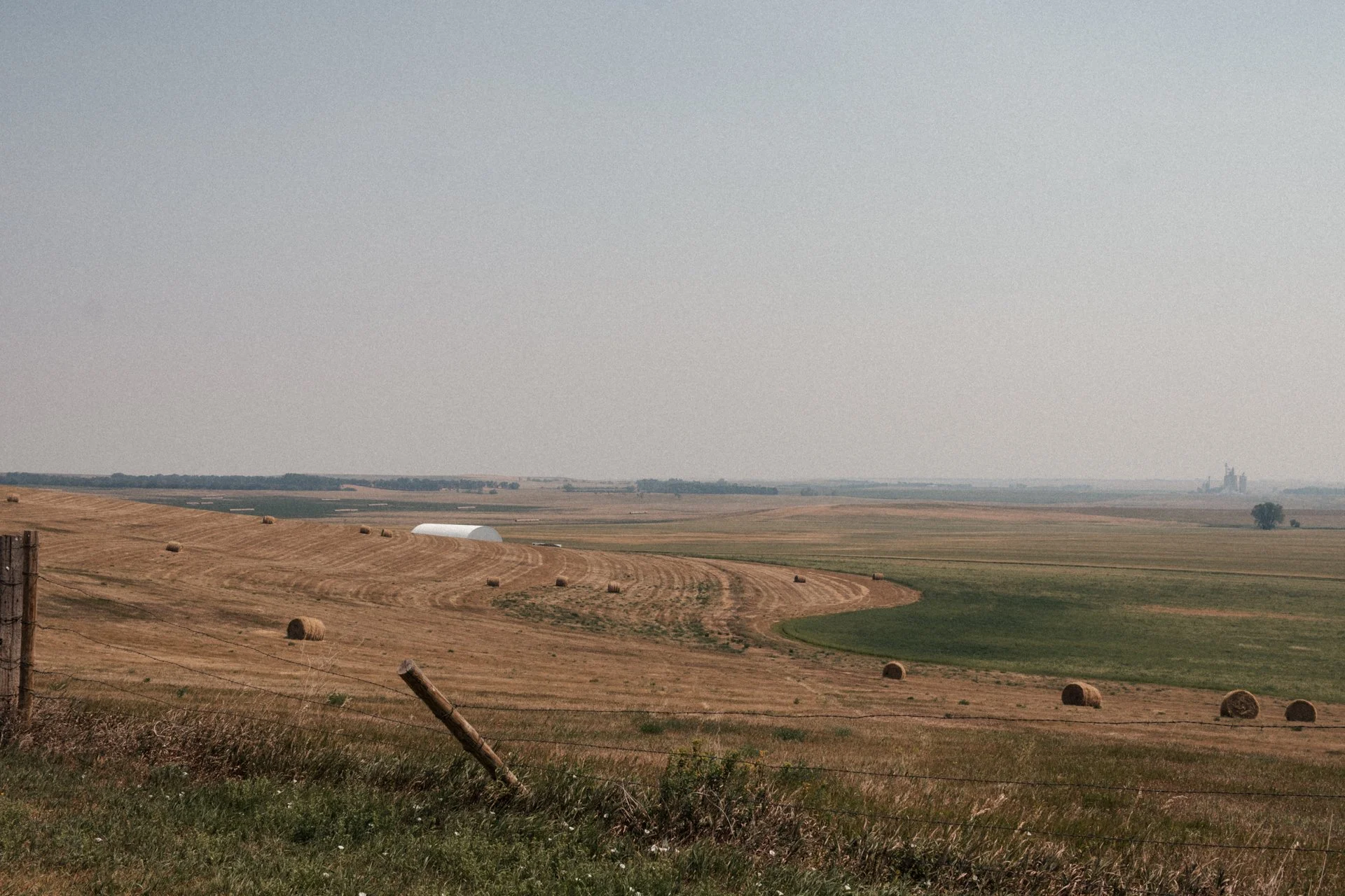 Open farmland with hay bales, a white barn, and distant silos under a gray sky.