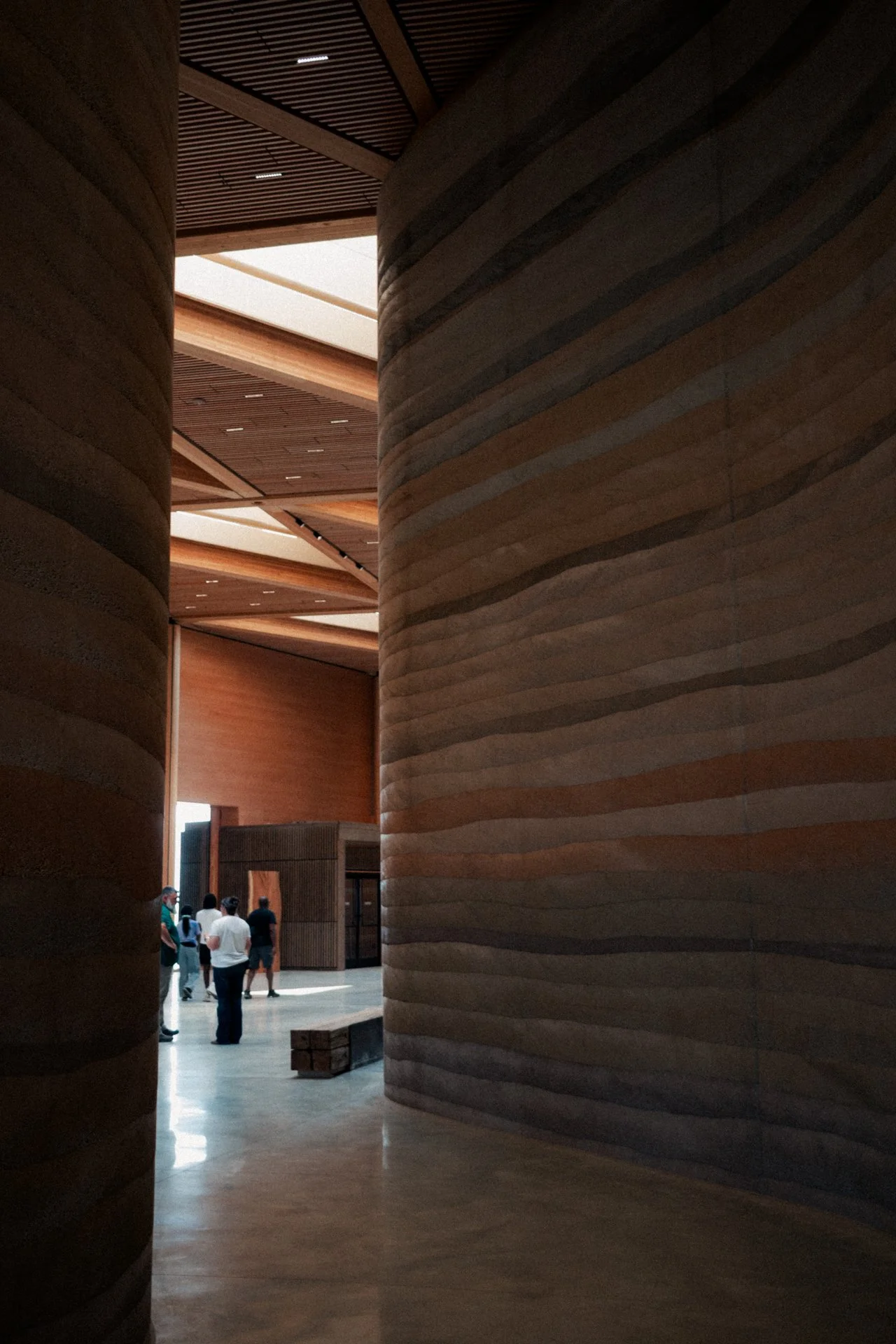 Interior view of a modern building with large wooden columns and a high ceiling, where a group of people are standing near an entrance or exhibit area.