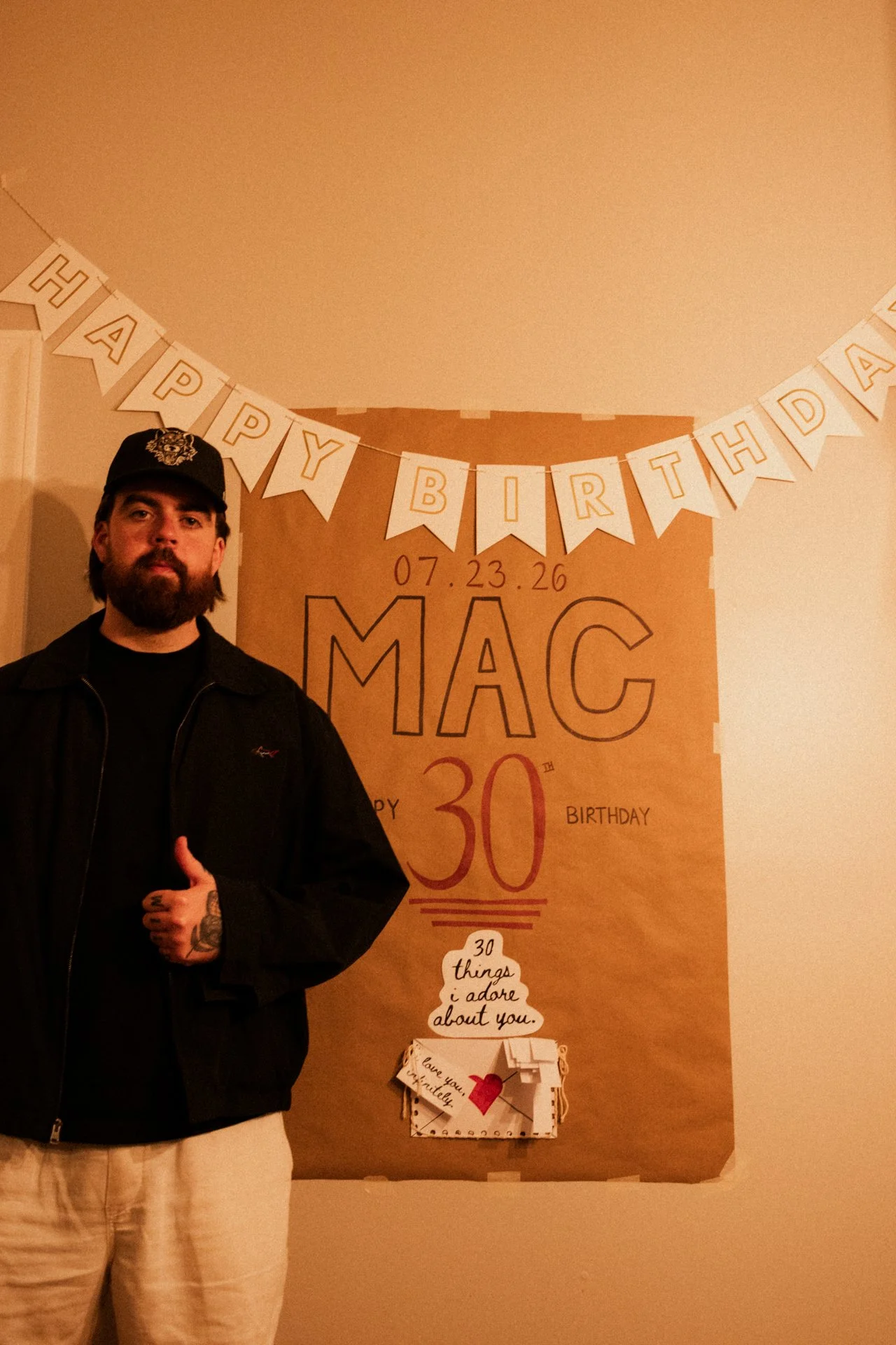 A man with a beard and tattoos giving a thumbs-up gesture, standing in front of a birthday decoration with a banner that says 'Happy Birthday' and a poster referencing '30 things I adore about you.'