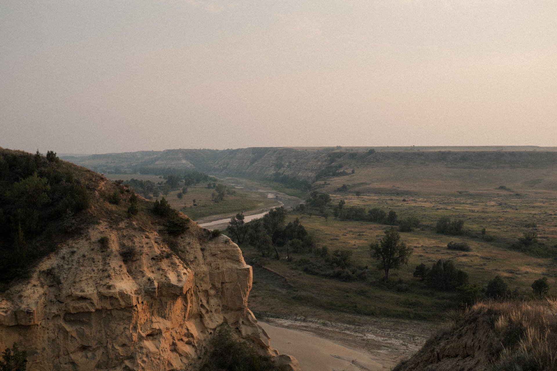 A scenic view of an open valley with rolling hills, a winding dirt road, sparse trees, and a distant flat-topped mesa under a hazy sky.
