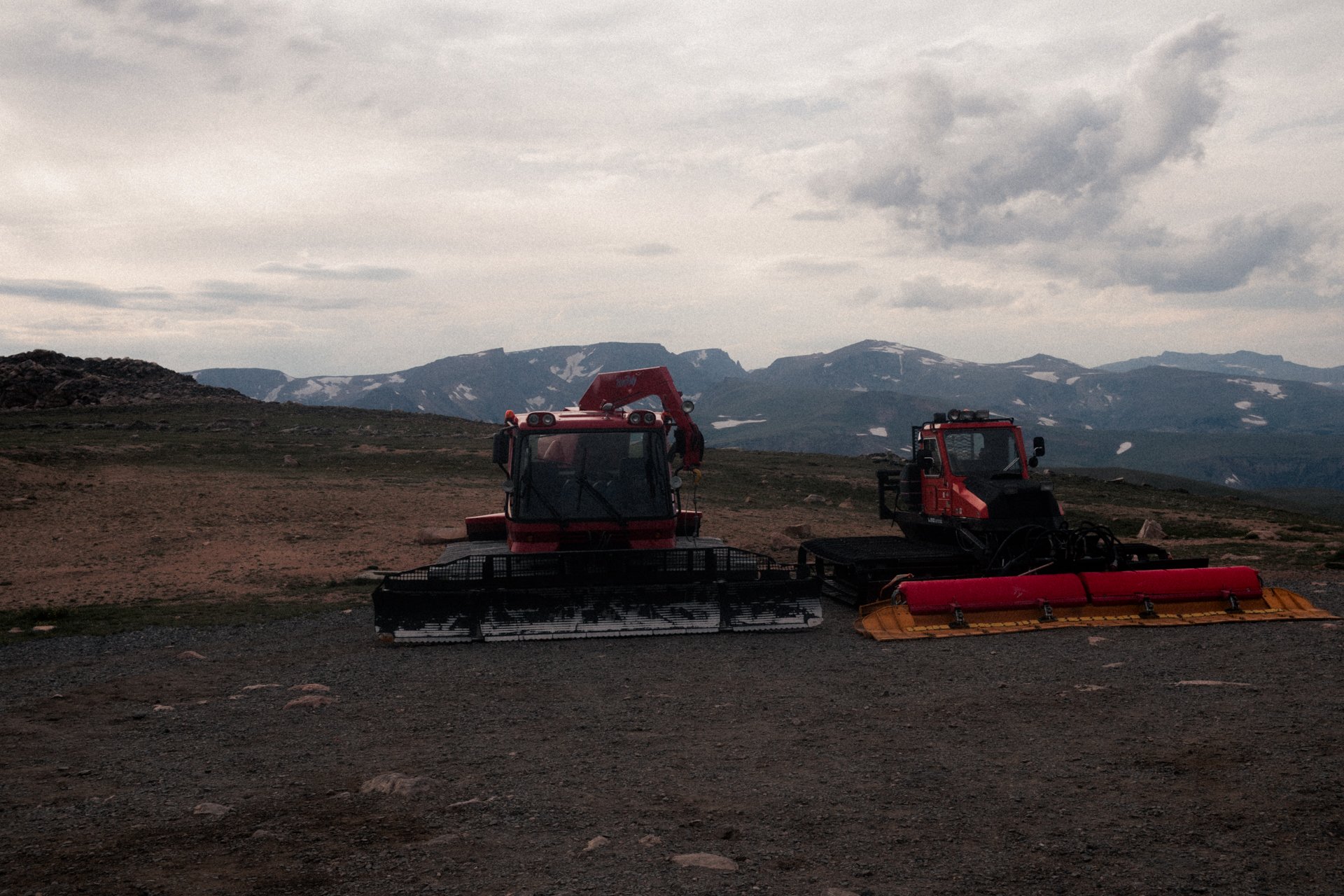 Two red snow grooming machines on a mountain landscape with snow patches, rocky terrain, and cloudy sky.