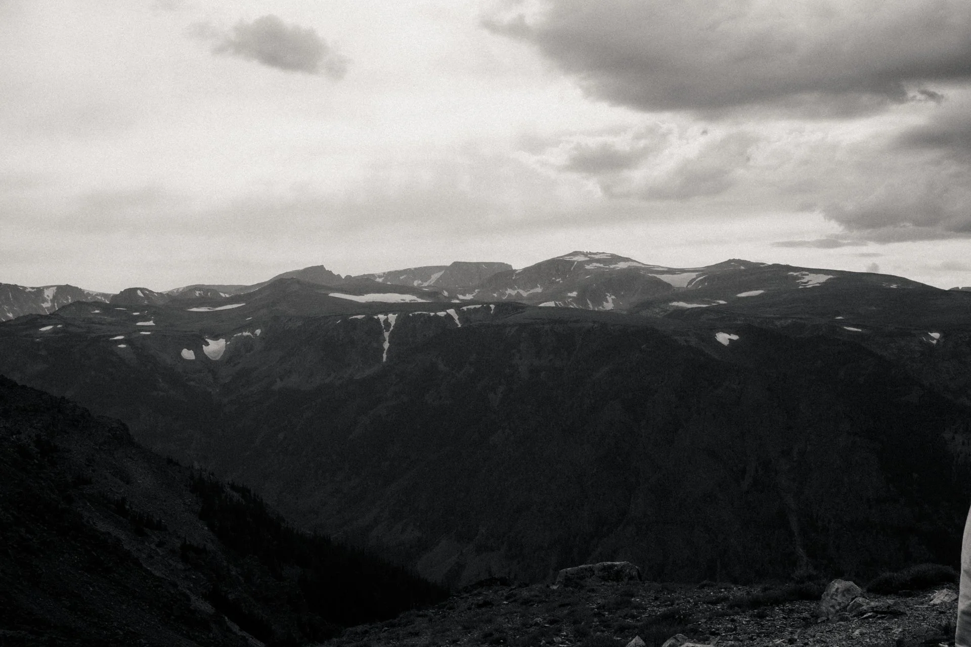 Black and white photo of a mountain range with patches of snow on the peaks, under a cloudy sky.