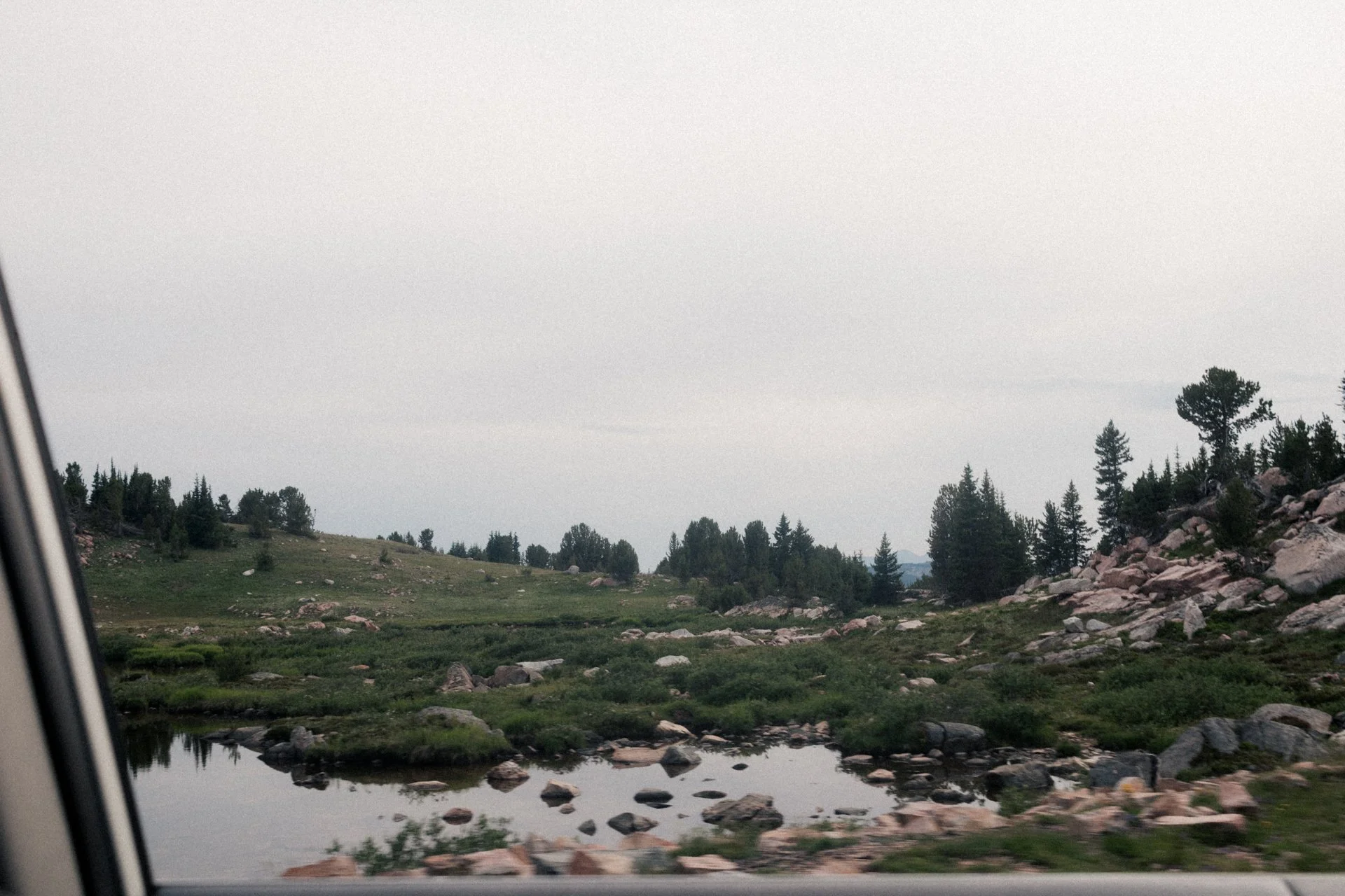 Scenic view of a lake and mountain landscape with green trees and rocky terrain, partly visible from a car window.