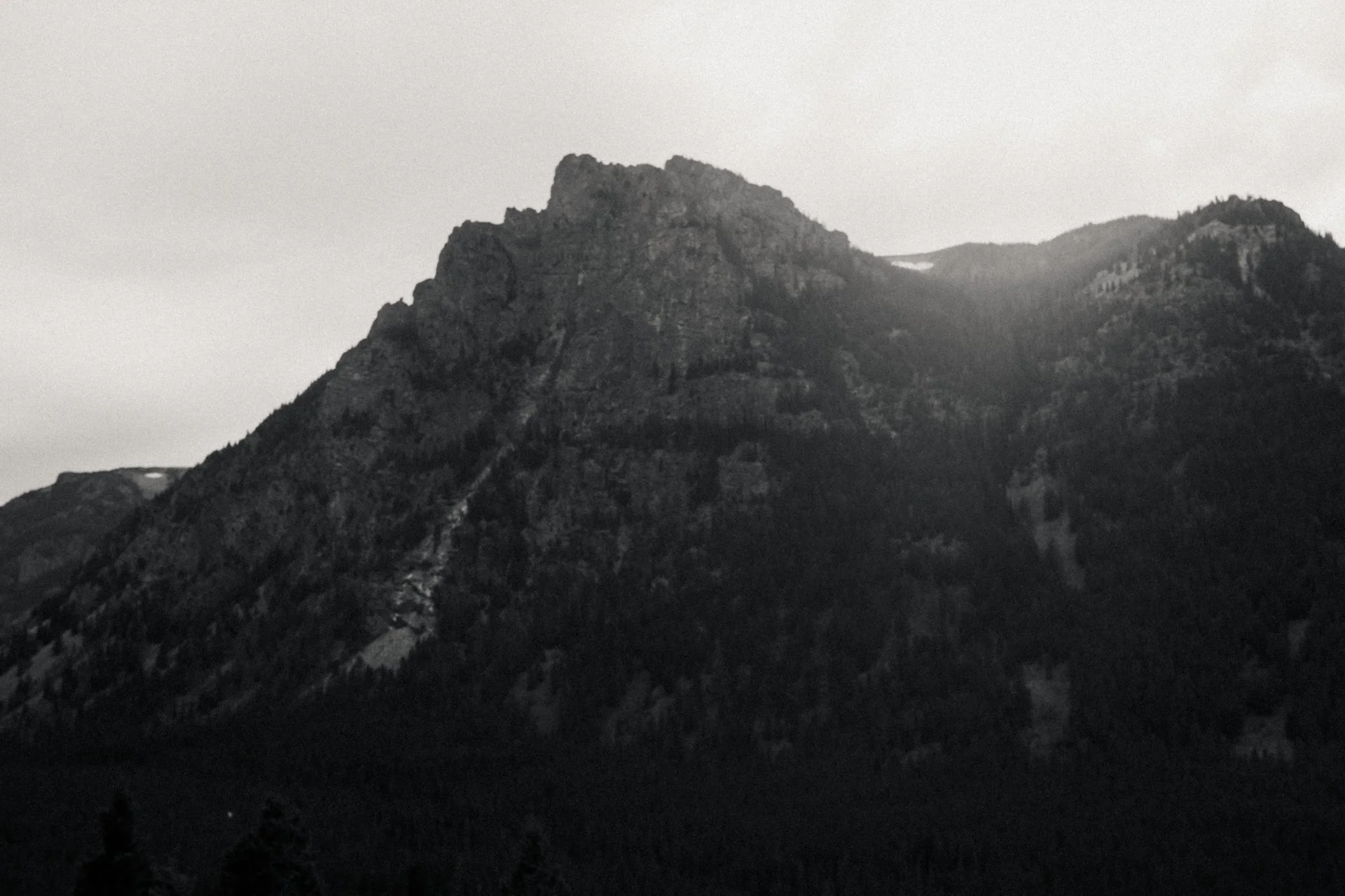 A black and white photo of a rugged mountain range with steep, jagged peaks and forested slopes.