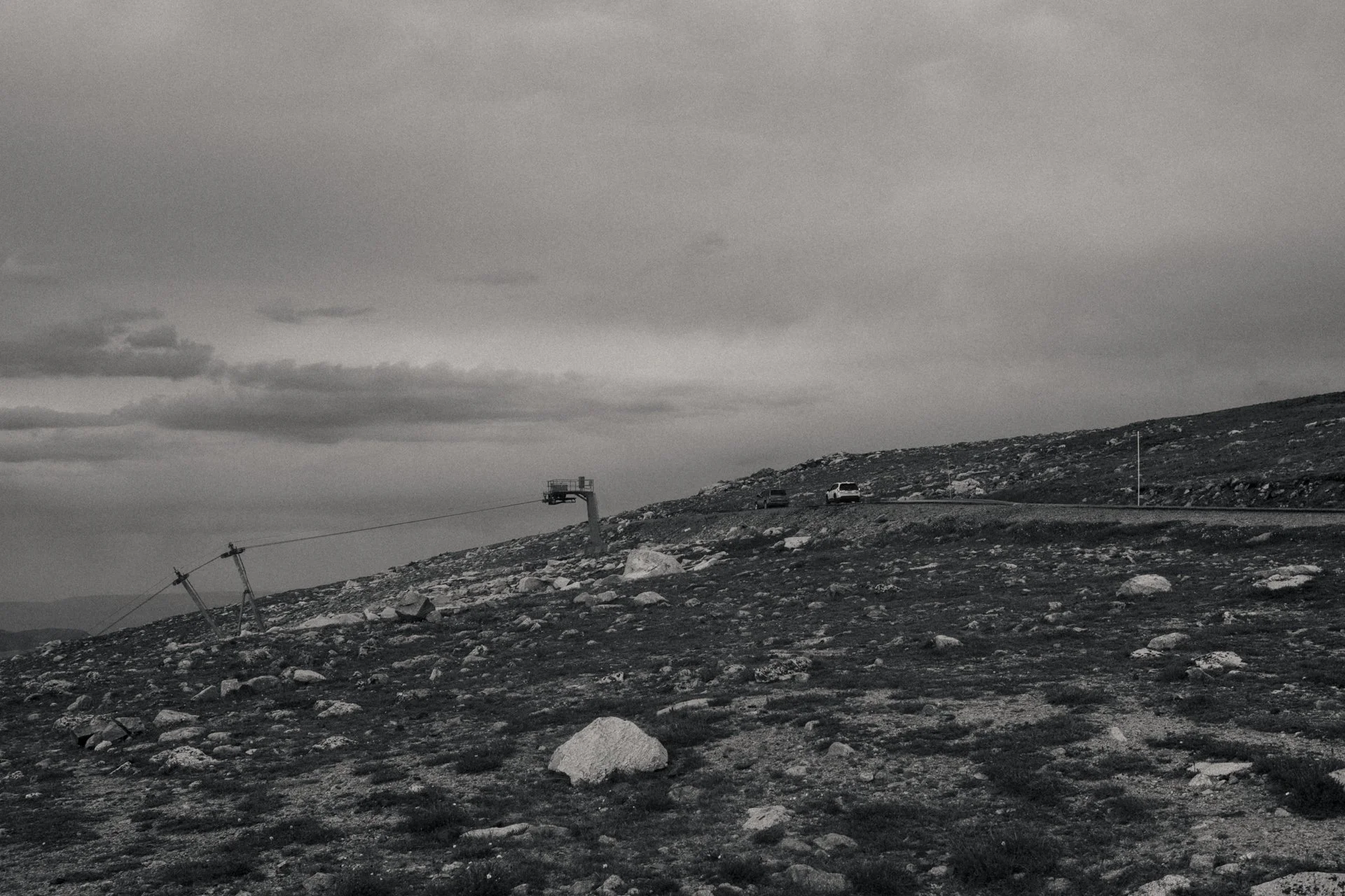Black and white photo of a mountainous landscape with rocky terrain, a chairlift, and a few parked vehicles against a cloudy sky.