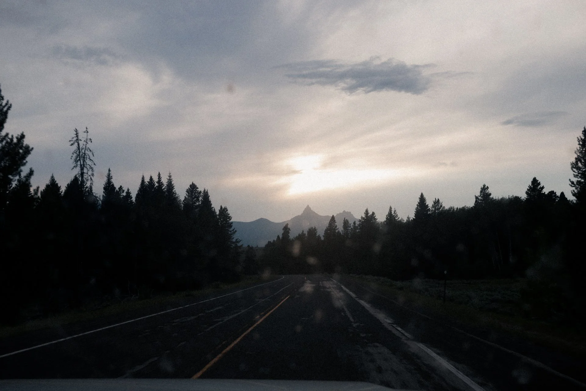 A scenic view of a mountain range partially covered by clouds, with the sun setting behind the mountains. The road in the foreground curves through a dense forest of tall pine trees.