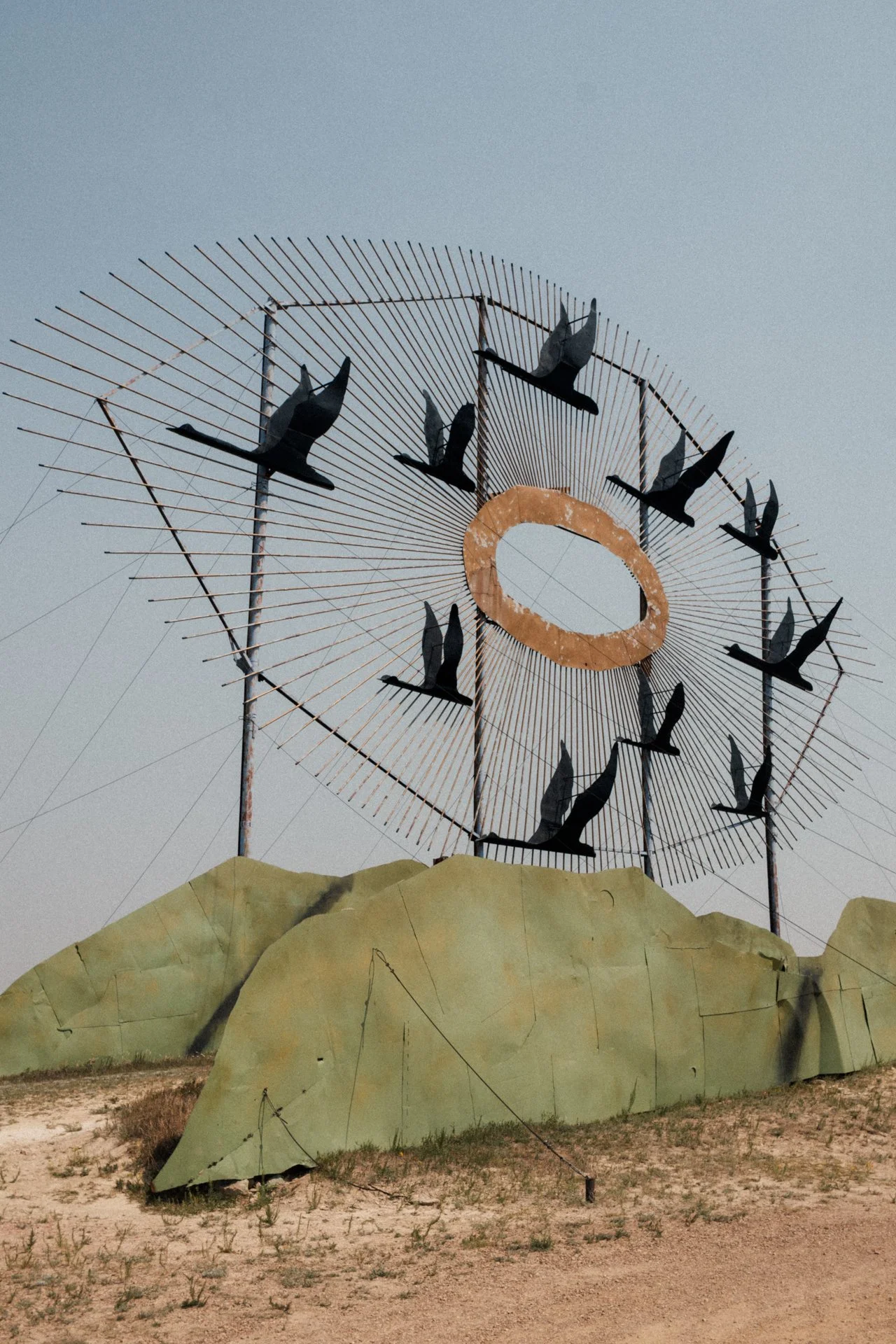 An outdoor art installation featuring a large circular metal structure with flying bird sculptures and a ring at the center, set against a cloudy sky and surrounded by a sandy ground with some grass.