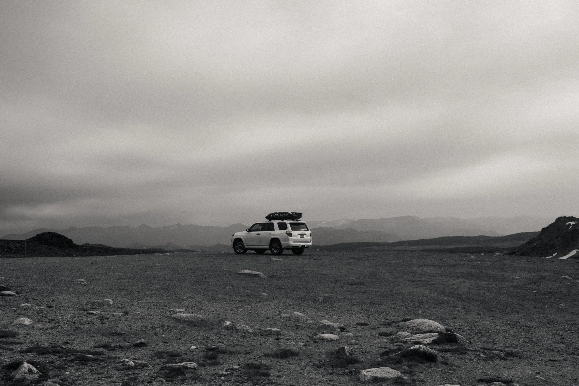 A white SUV with a roof cargo box parked on a barren, rocky landscape under a cloudy sky, with distant mountains on the horizon.