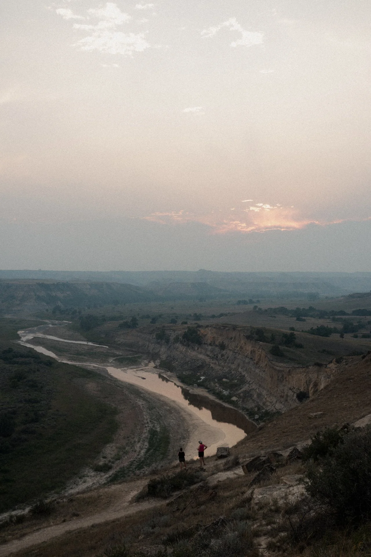 Two people walking along a dirt trail overlooking a winding river in a valley during sunset, with hills and sparse vegetation.