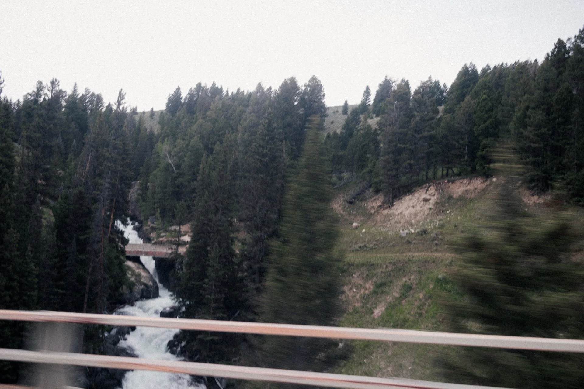 Scenic view of a forested mountain landscape with a waterfall and a bridge, viewed from a moving vehicle.