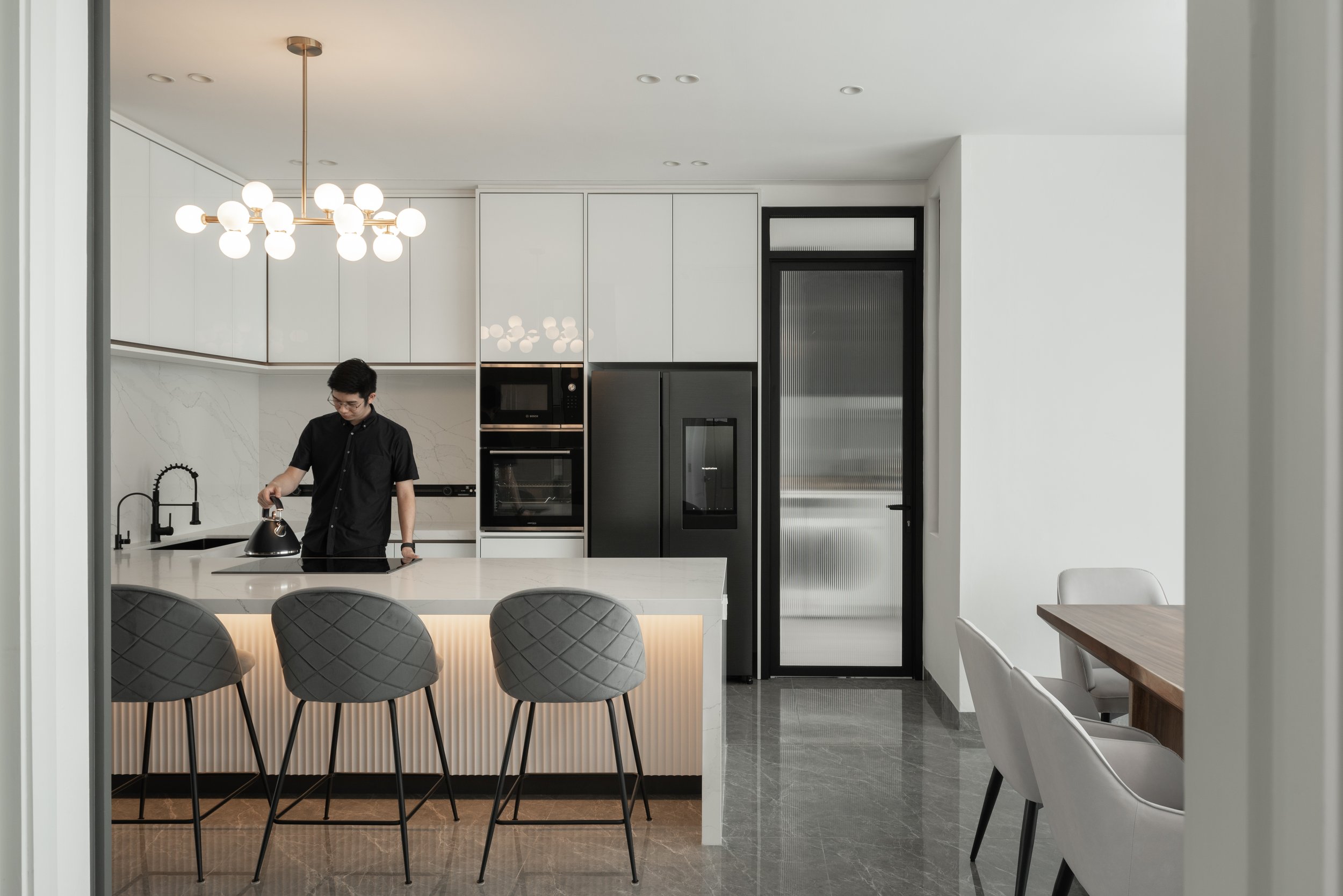 Modern kitchen with a man preparing a beverage at the counter, black and white cabinets, black refrigerator, and a dining table with white chairs.