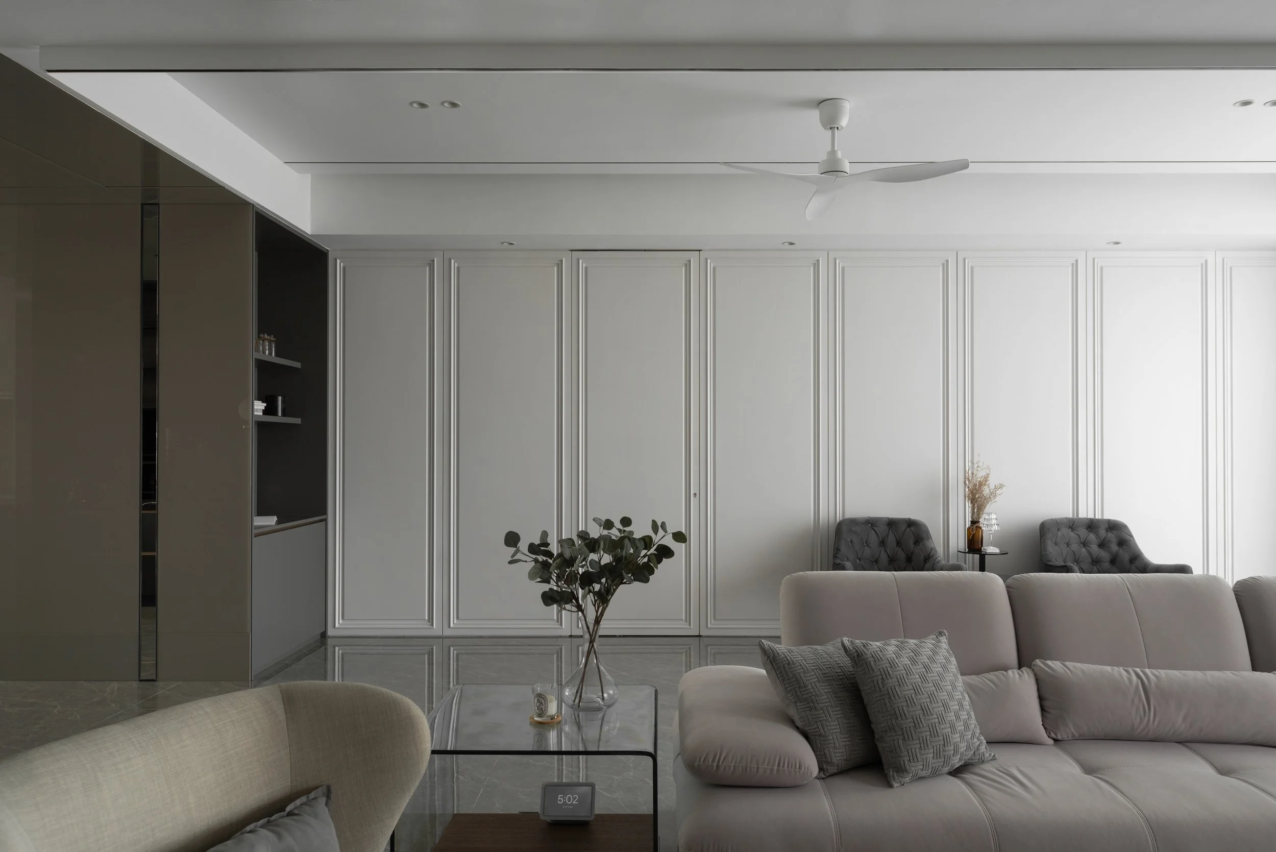 Minimalist living room with white walls, a ceiling fan, gray and beige sofas, a glass coffee table with a vase of greenery, and decorative pillows.