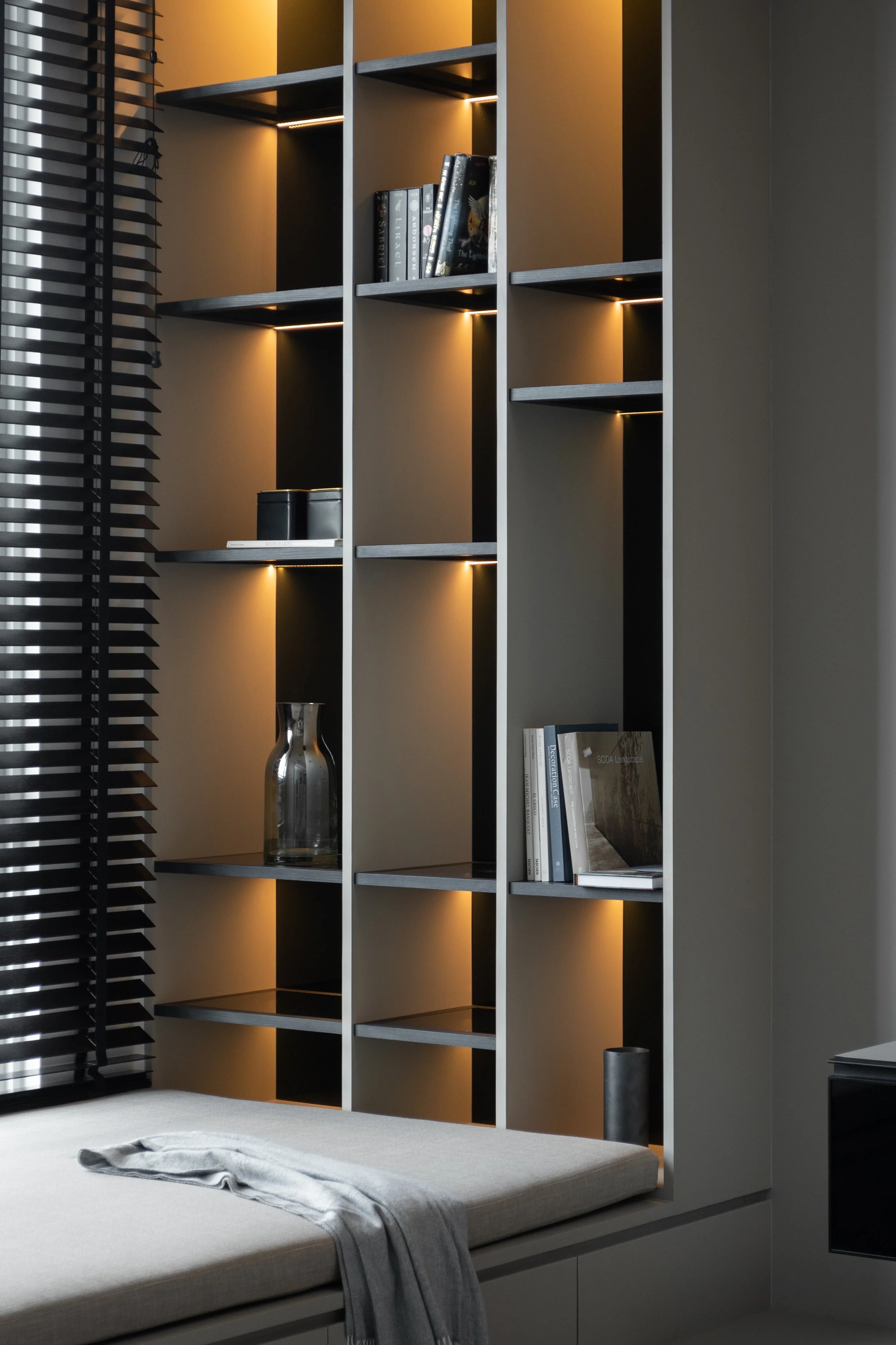 Modern bookshelf with built-in lighting, containing books and decorative items, adjacent to a window with horizontal blinds and a beige cushioned bench with a gray throw blanket.