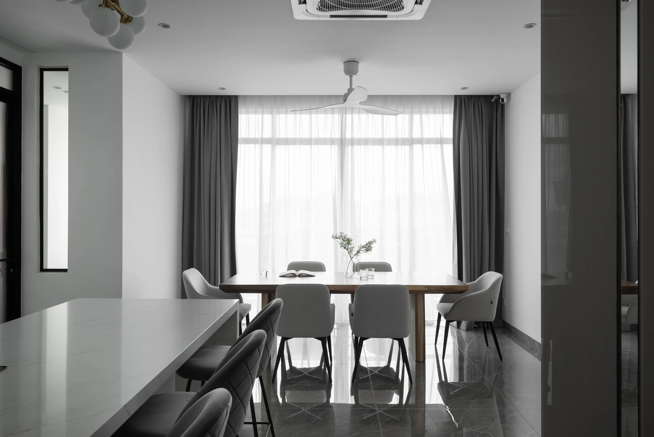 Modern dining room with a wooden table, six upholstered chairs, large windows with light sheer and dark curtains, ceiling fan, and ceiling light fixture.