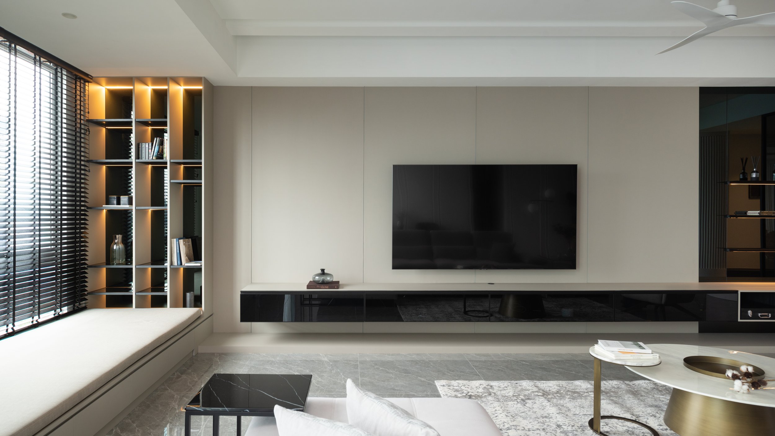 Modern living room with built-in shelves on the left, a large flat-screen TV on the wall, and a window seat with black blinds.