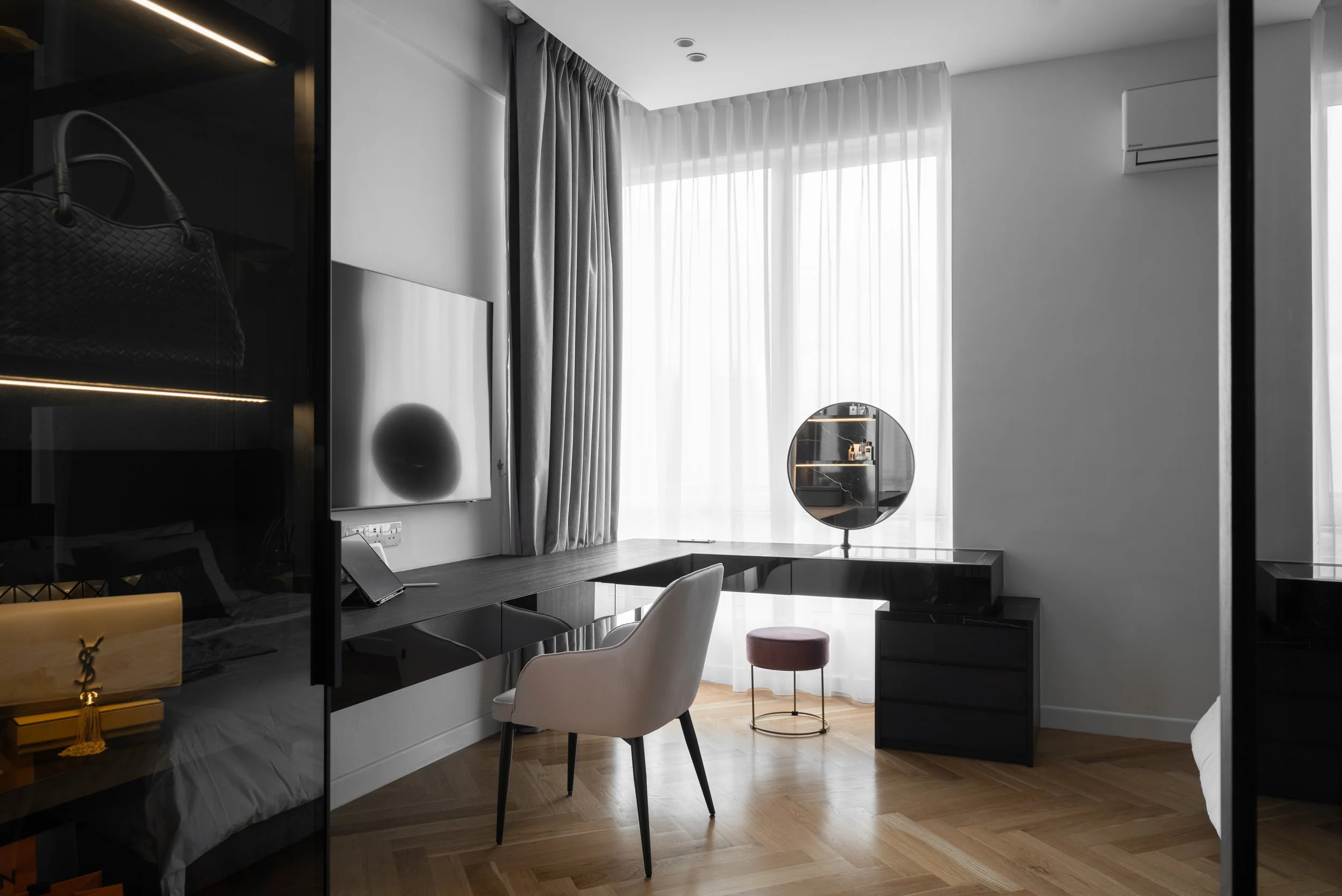 Modern hotel room with a large window covered by sheer curtains and heavy drapes, a flat-screen TV mounted on the wall, a white upholstered chair at a sleek black desk, a round pink stool, and a black vanity table with a mirror. The room features woo