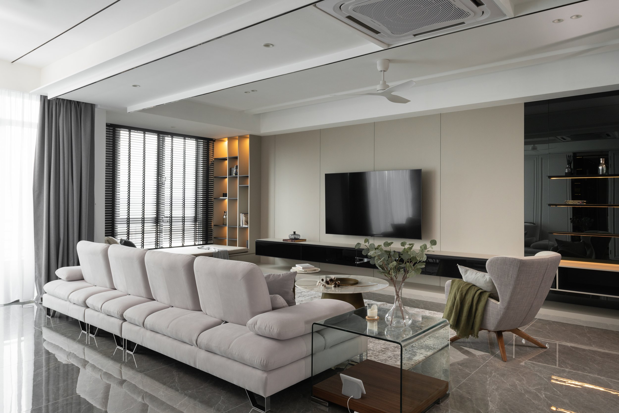 Modern living room with a large white sofa, a gray armchair with a green blanket, a round coffee table with decorative items, a flat-screen TV mounted on the wall, black shelving below, and a floor-to-ceiling window with blinds.
