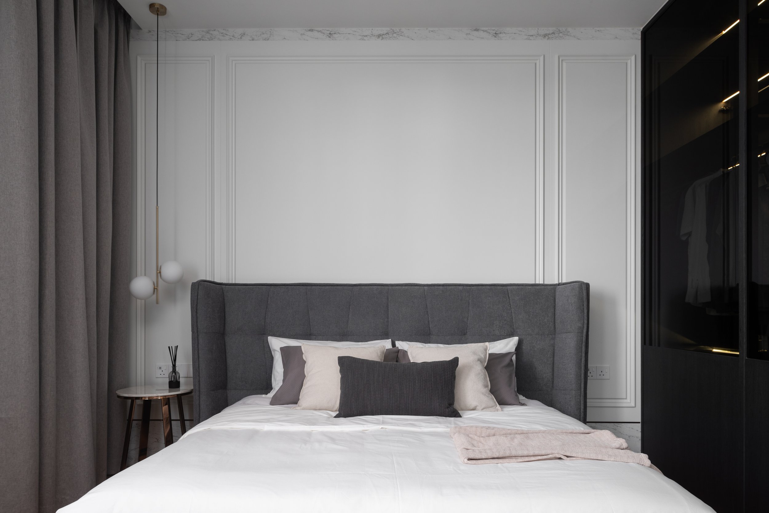 A modern bedroom with a large bed featuring gray and white pillows, a dark gray upholstered headboard, a small round side table with a black reed diffuser, dark gray curtains, and a black wardrobe with warm interior lighting.