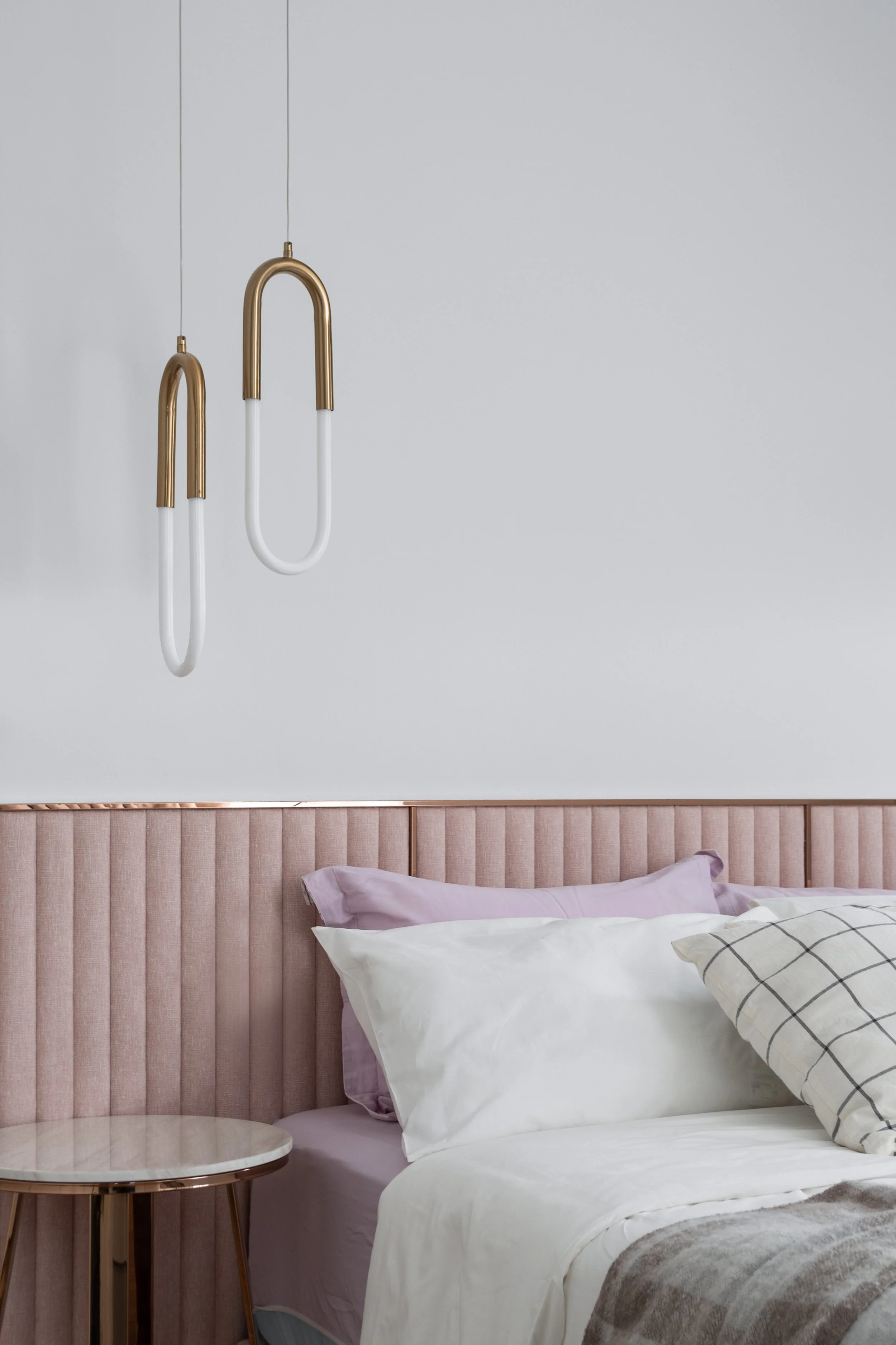 Modern bedroom with a pink upholstered headboard, white and plaid pillows, a beige blanket, a small round side table, and two hanging pendant lights with gold and white design against a light gray wall.