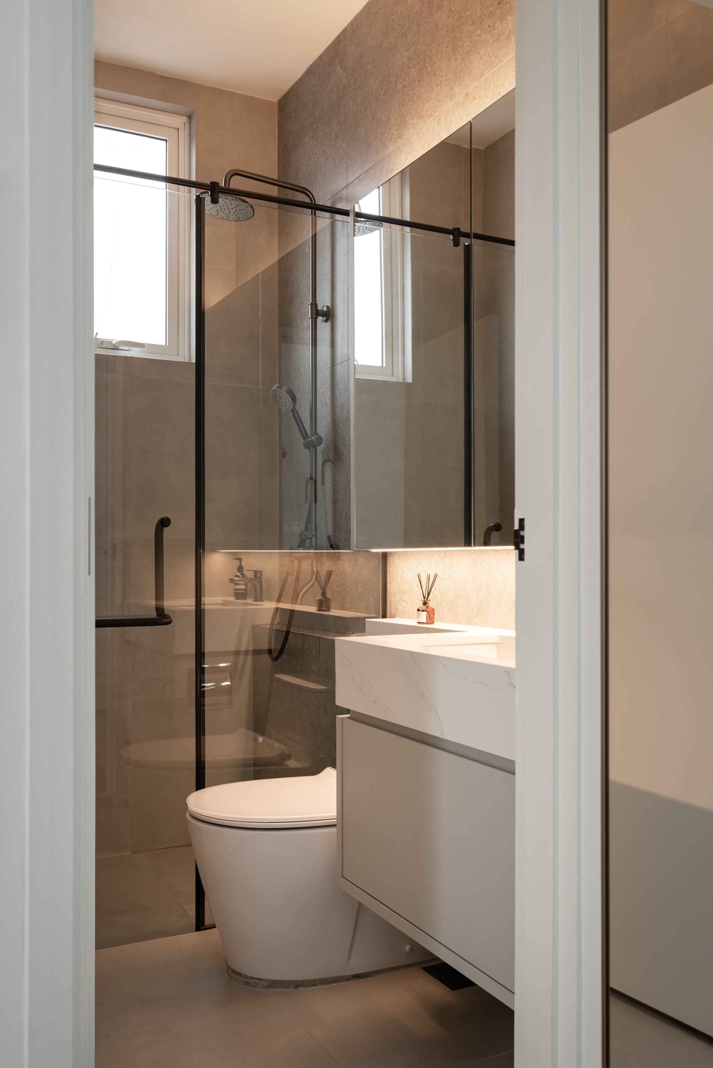 Modern bathroom with a glass shower enclosure, a white toilet, and a vanity with a marble countertop and reed diffuser, illuminated by natural light from a window.