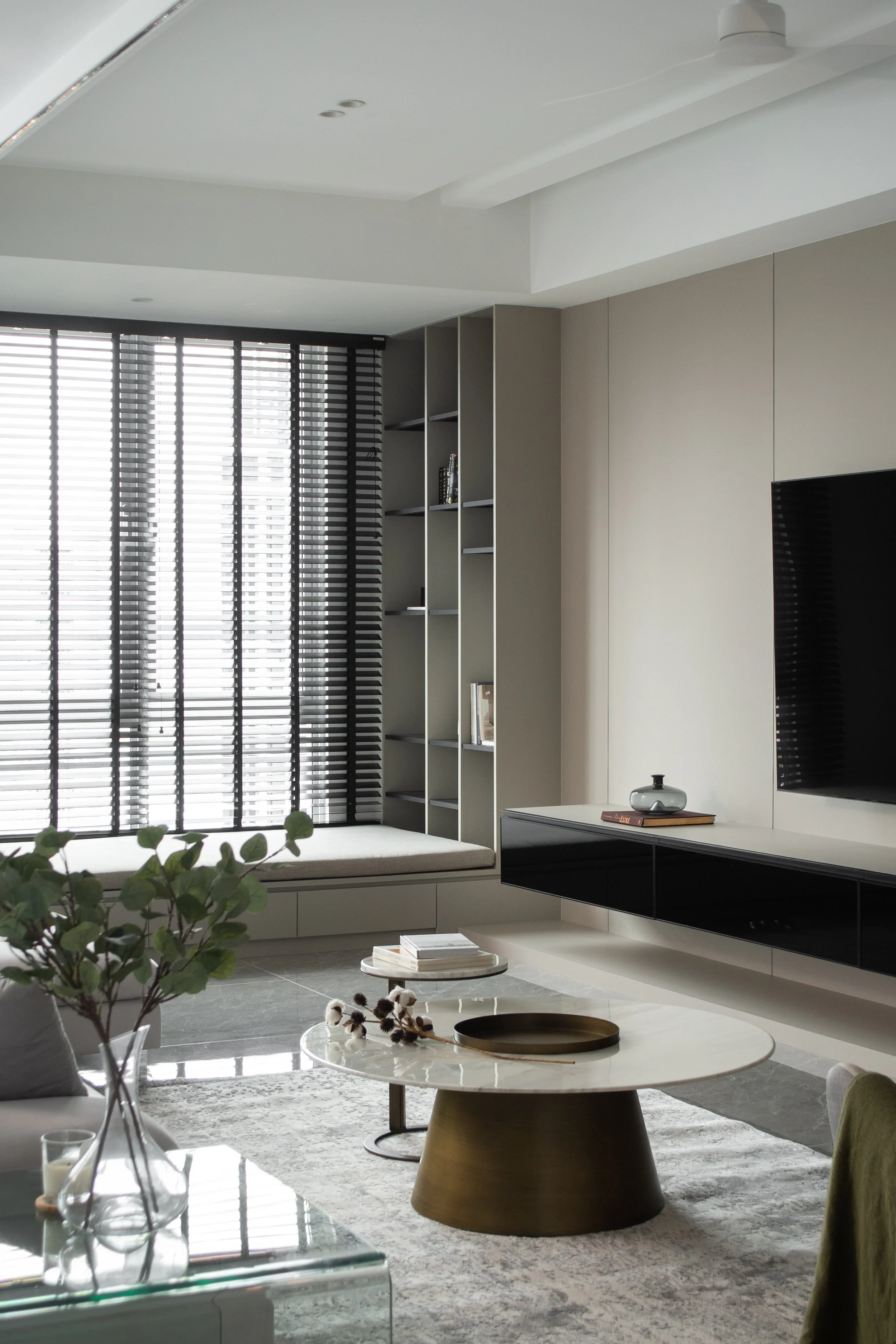 Modern living room with a large window with black blinds, neutral-colored walls, a built-in bench under the window, a wall-mounted flat-screen TV, a coffee table with a tray and decorative items, and a bookshelf with books and decor.
