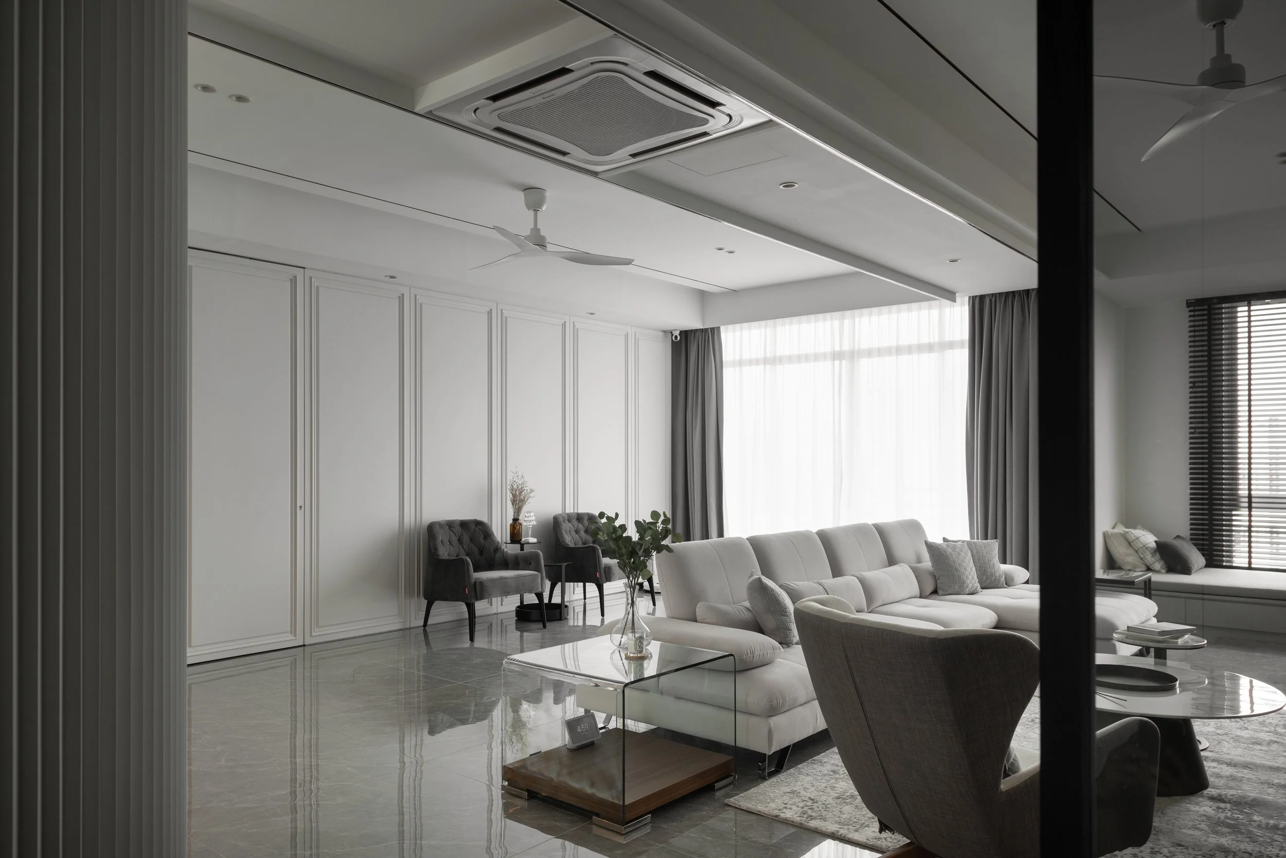 Modern living room with white walls, large window with sheer curtains, white sectional sofa, armchair, glass coffee table, and decorative plants.