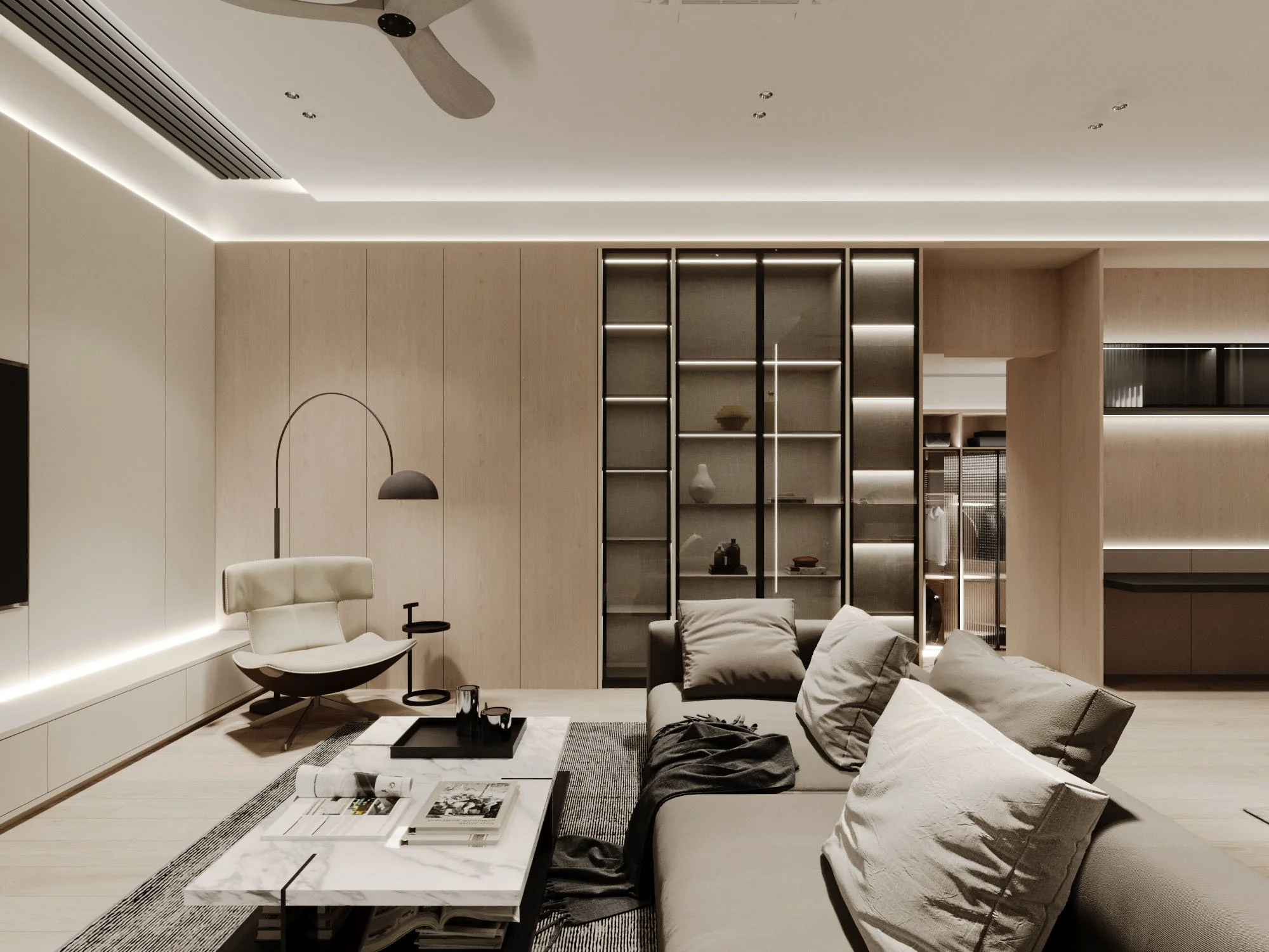 Modern living room with beige walls, a gray sofa with pillows, a coffee table with magazines, a white armchair with a black side table, and a black and white shelving unit with decorative items. Recessed lighting and a ceiling fan are visible.