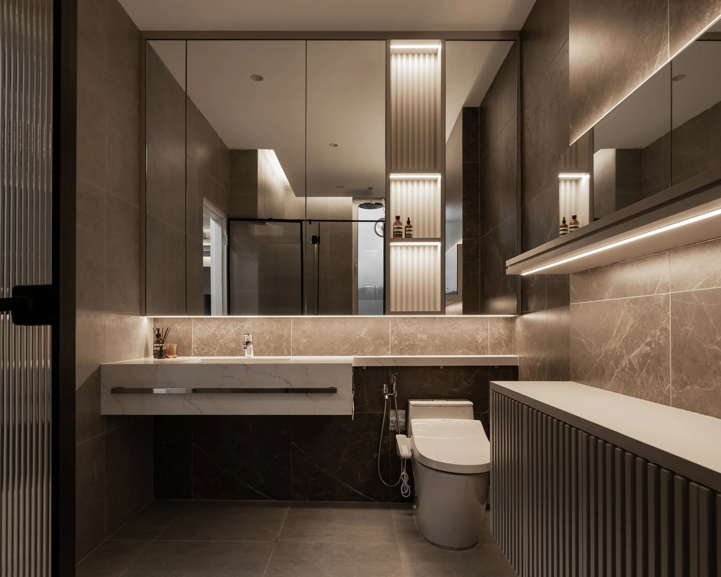 Modern bathroom with a large mirror, marble countertops, and sleek cabinetry. Features include a white toilet, a small sink, and decorative bottles on shelves. The lighting is soft and ambient.