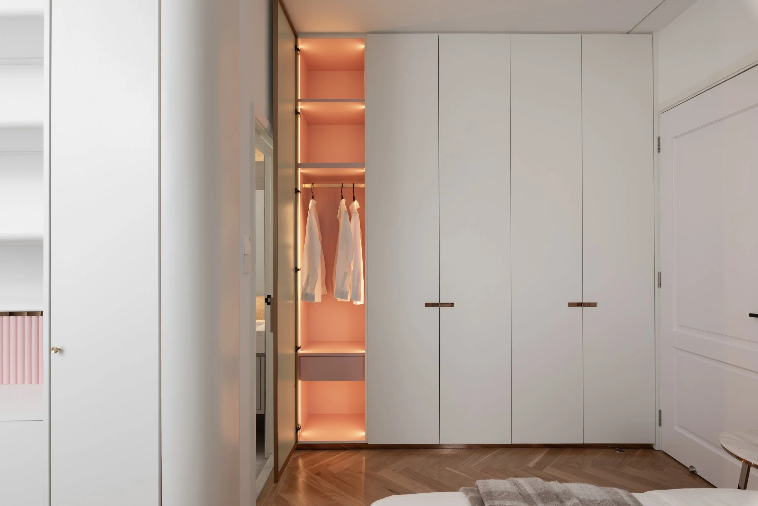 Interior view of a bedroom with a closed white wardrobe, an open closet with warm lighting and hanging clothes, and a herringbone wood floor.