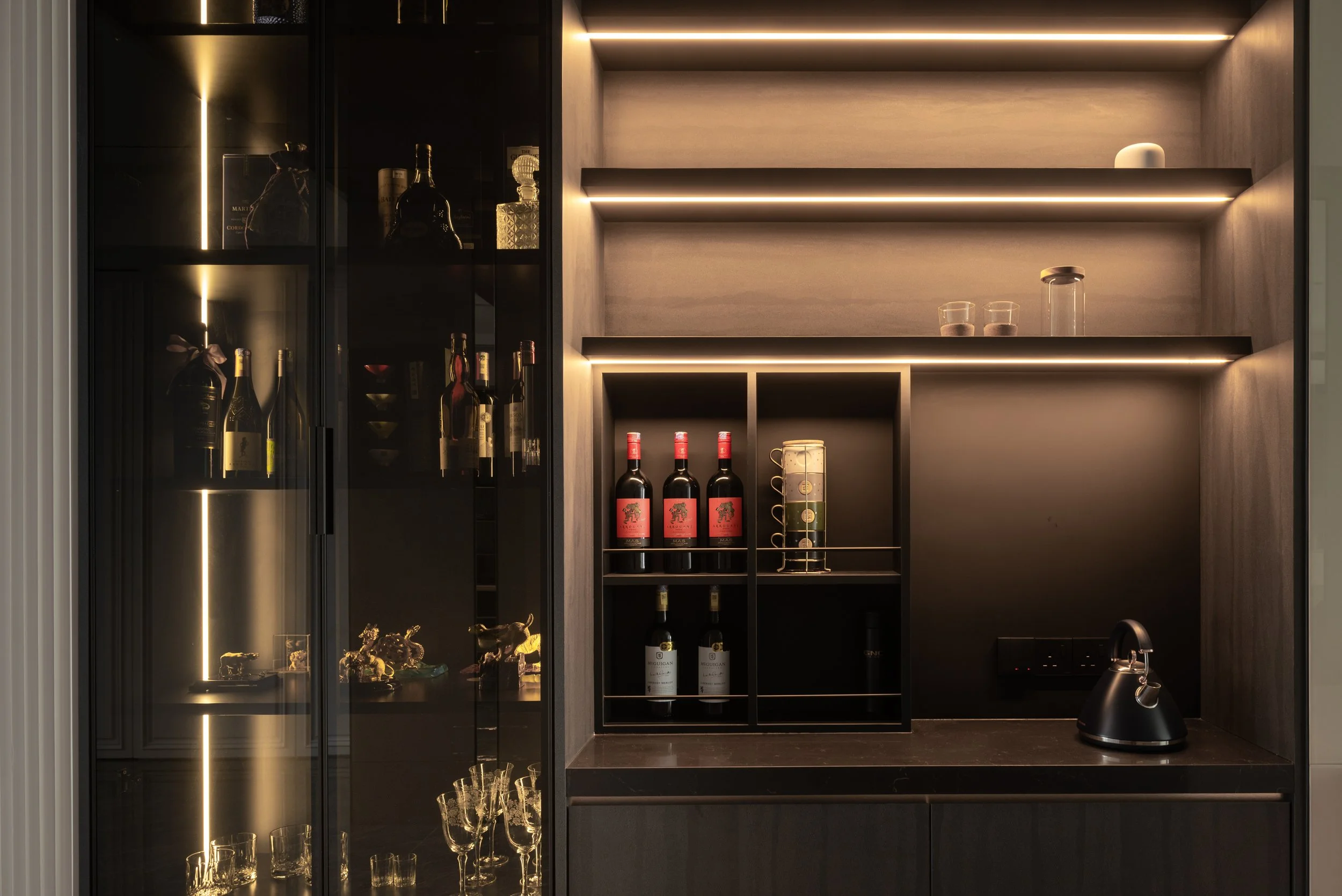 Modern home bar area with glass-front cabinets containing liquor bottles and glassware, and a dark countertop with a black kettle.