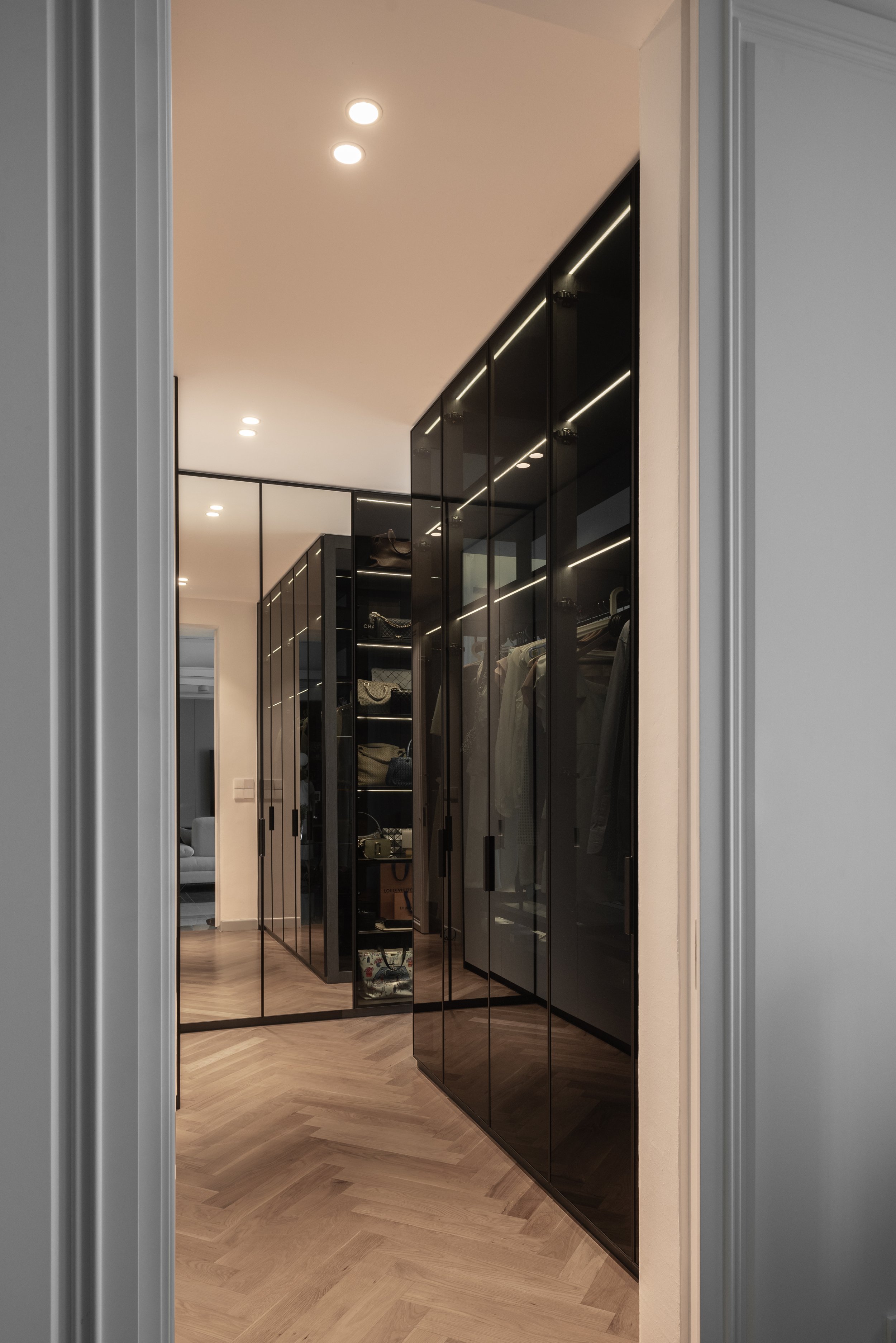 A modern walk-in closet with black glass doors and built-in shelving for clothing and accessories, illuminated by recessed ceiling lights.